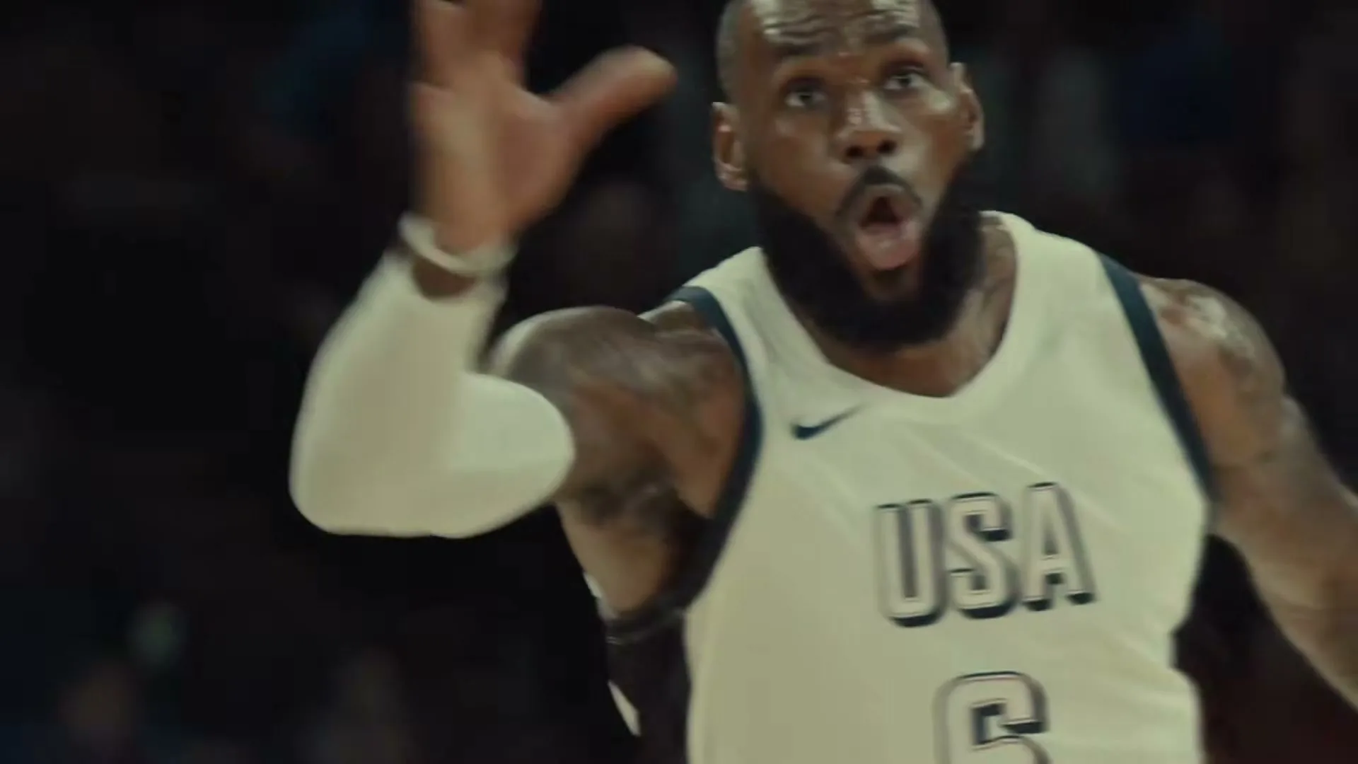 LeBron James, a male basketball player in his 30s, wearing a white USA jersey, looks surprised and gestures with his hand on a basketball court.