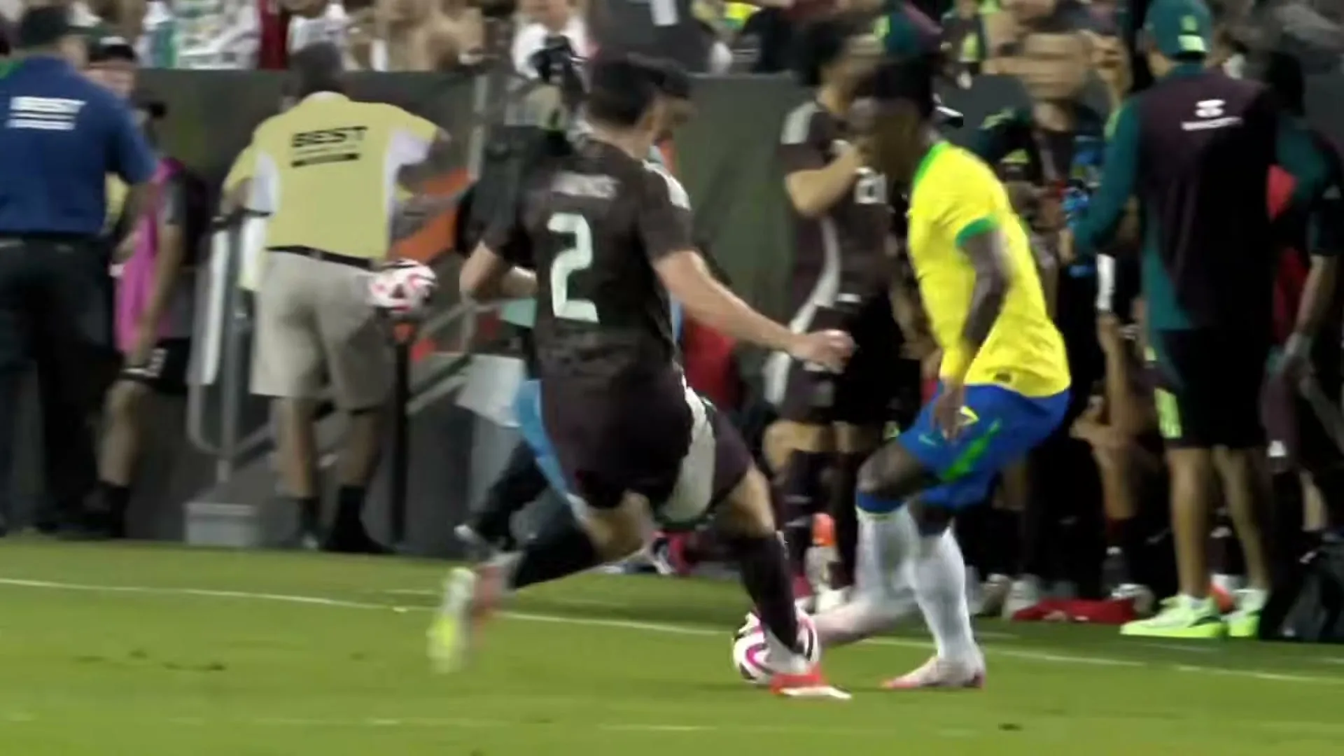 Two male soccer players, one in a black jersey and one in a yellow jersey, compete for the ball on a green field during a game.