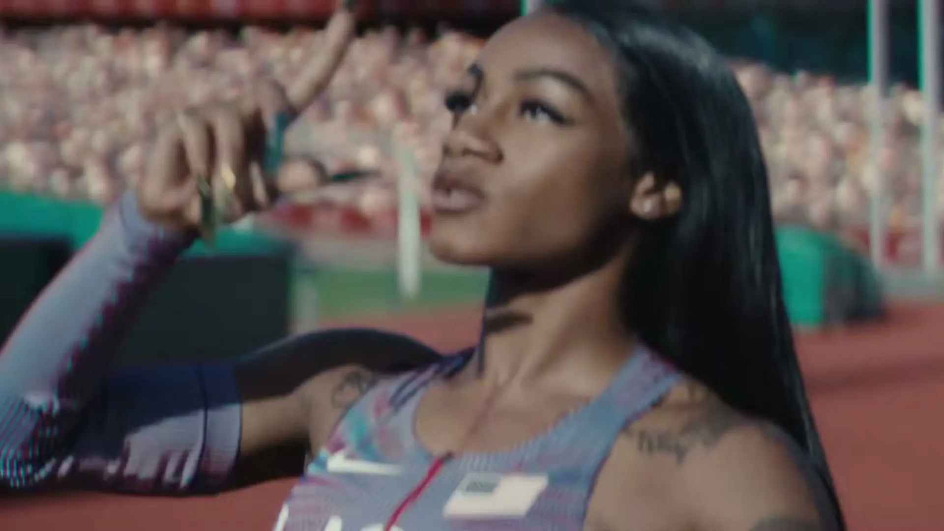 A female athlete with long dark hair, wearing a USA track uniform, looks up and adjusts her hair on a track field with stadium seating in the background.