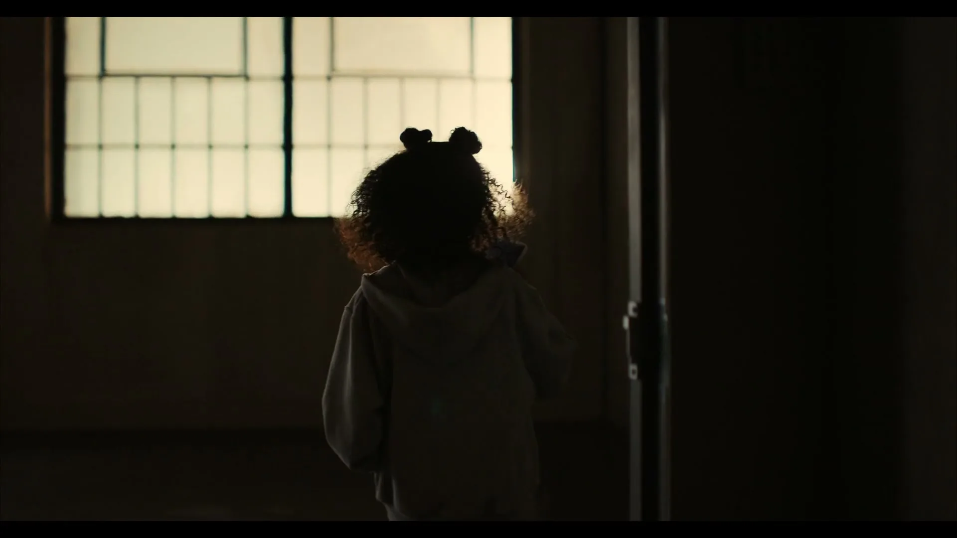 A young girl with curly hair and two buns walks away from the camera into a dimly lit room with a window, holding a small object.