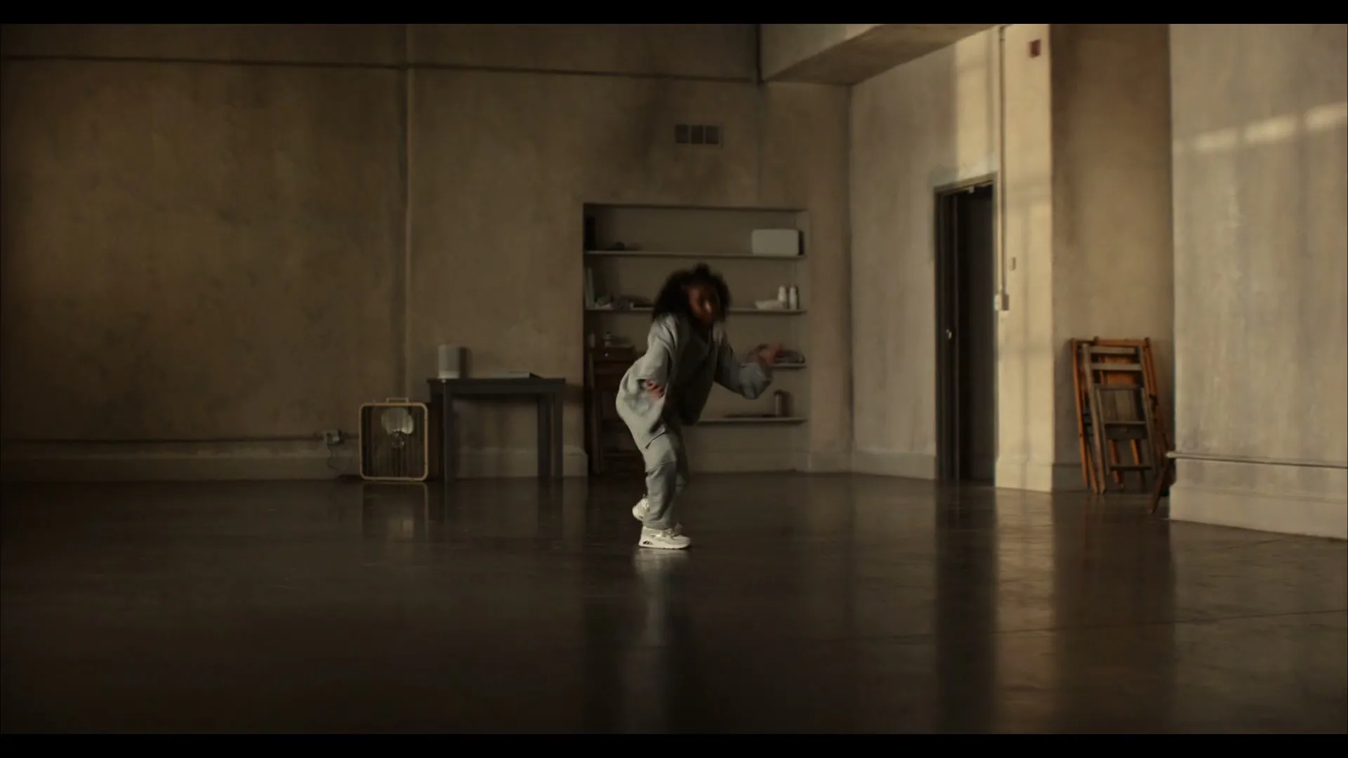A young female dancer with curly hair, wearing a grey tracksuit, practices her moves in a spacious, empty studio with concrete walls.