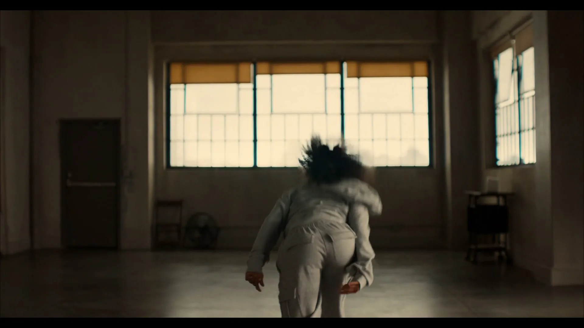 A young woman with curly hair, wearing a grey sweatsuit, dances energetically in a large, empty room with windows in the background.