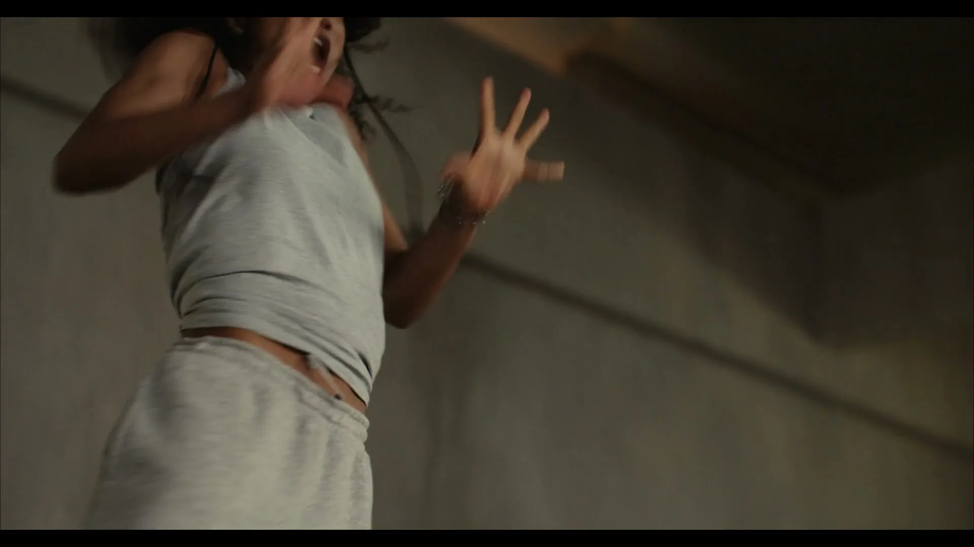 A young woman with dark, curly hair and a tank top jumps and screams with excitement in a dimly lit, industrial-style room.