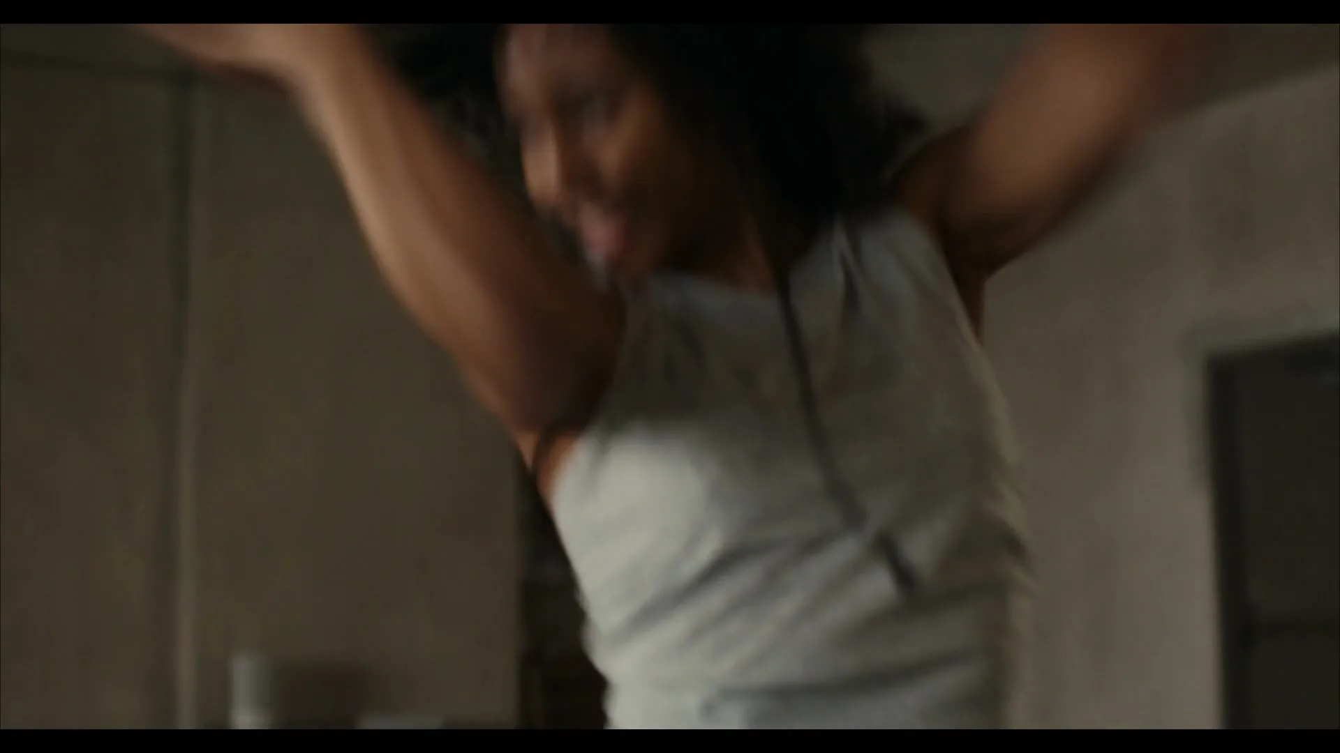 A young woman with curly hair, wearing a tank top, dances energetically in a sparsely furnished room, moving her arms and body to the music.
