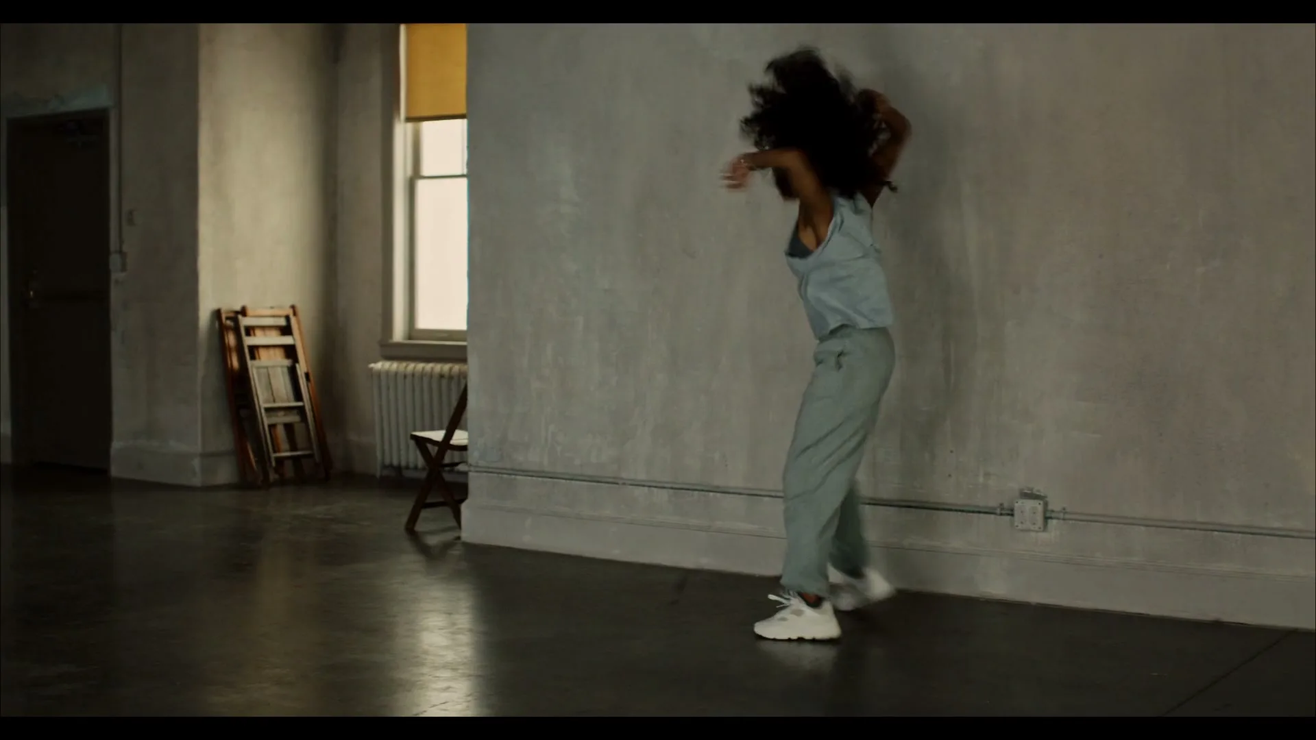 A young woman with curly hair, wearing a light blue tank top and grey sweatpants, dances energetically in a spacious, sparsely furnished room with concrete walls and a large window.