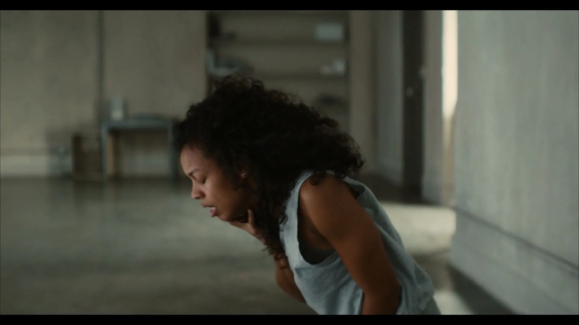 A young woman with curly hair, wearing a gray tank top and sweatpants, kneels on a concrete floor, looking down and holding her chest.