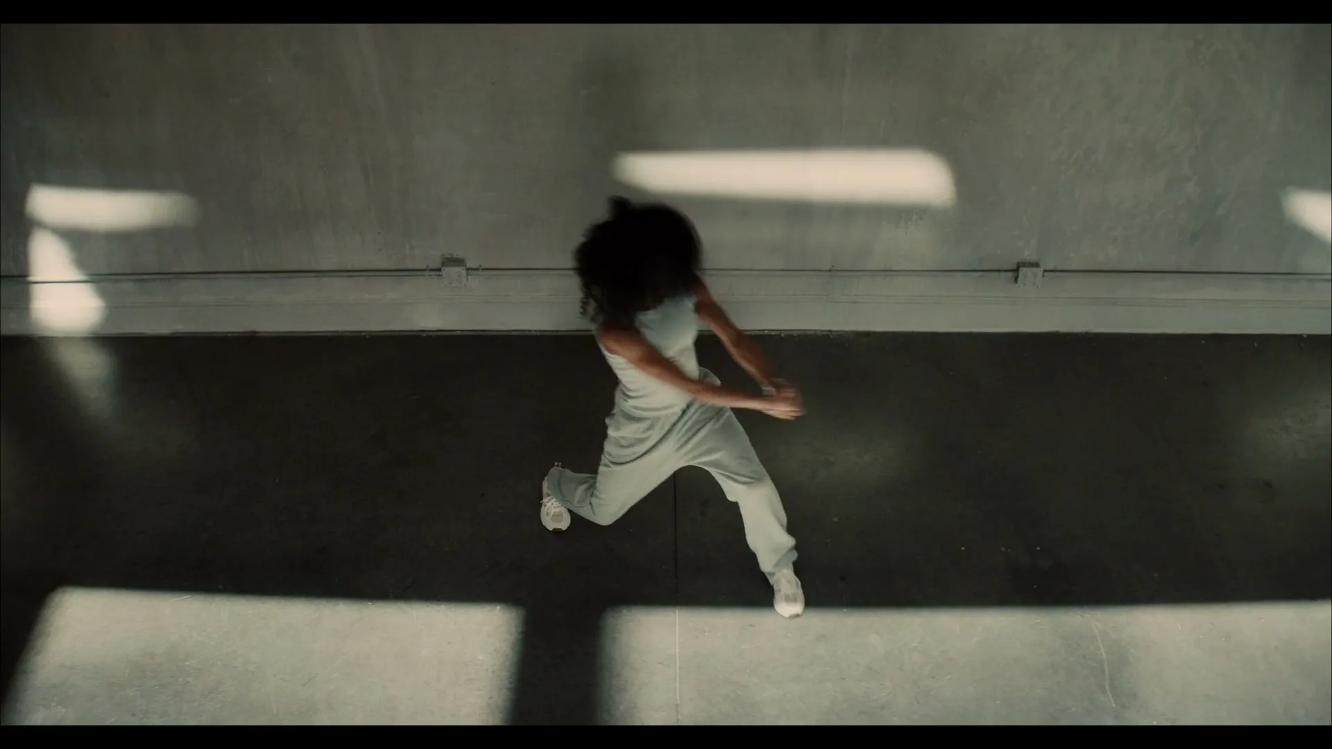 A female dancer with curly hair, wearing a light grey crop top and sweatpants, performs a contemporary dance routine on a concrete floor with sunlight streaming in.