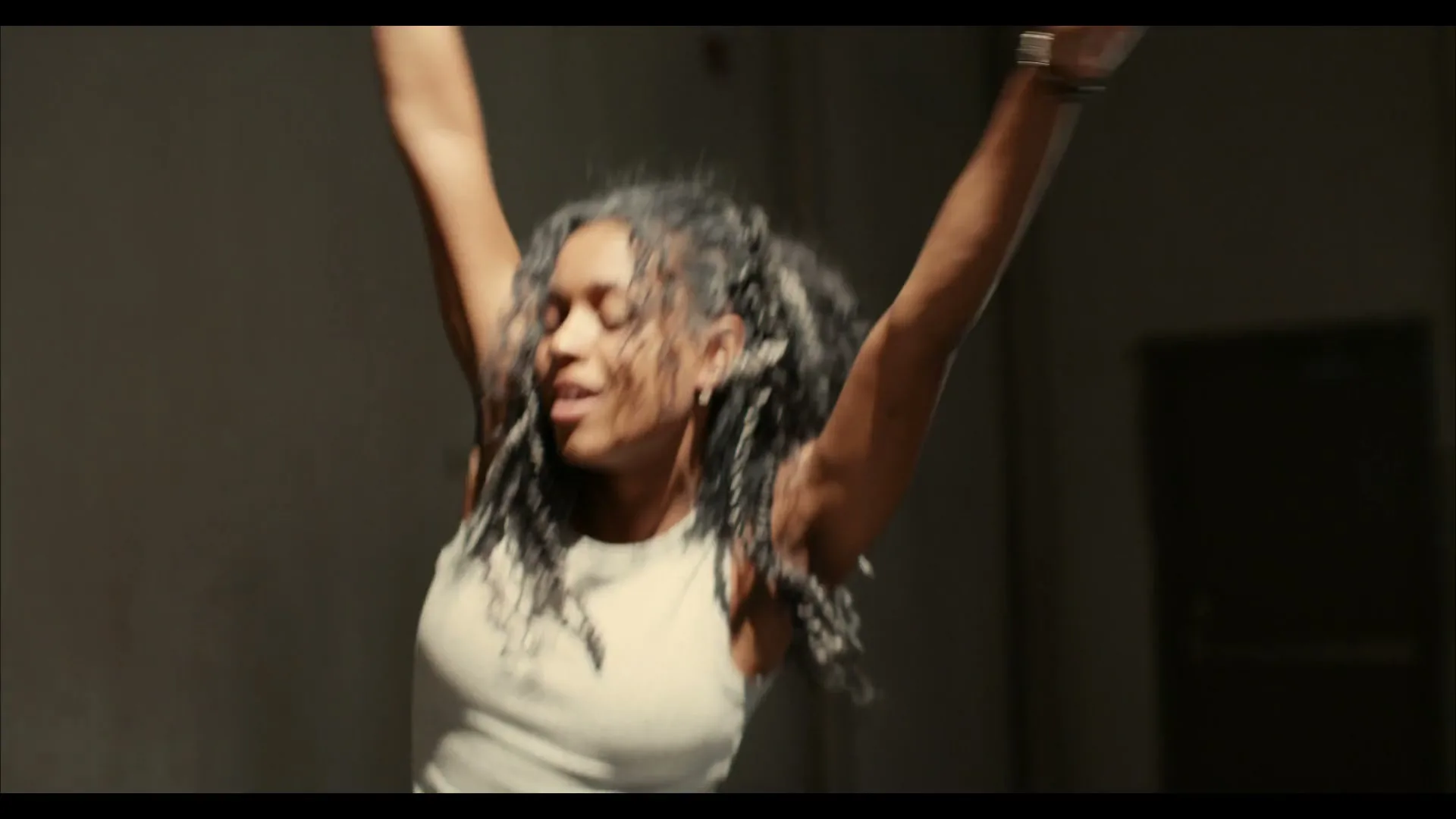 A young woman with braided hair, wearing a tank top, dances energetically with her arms raised in a dimly lit room.