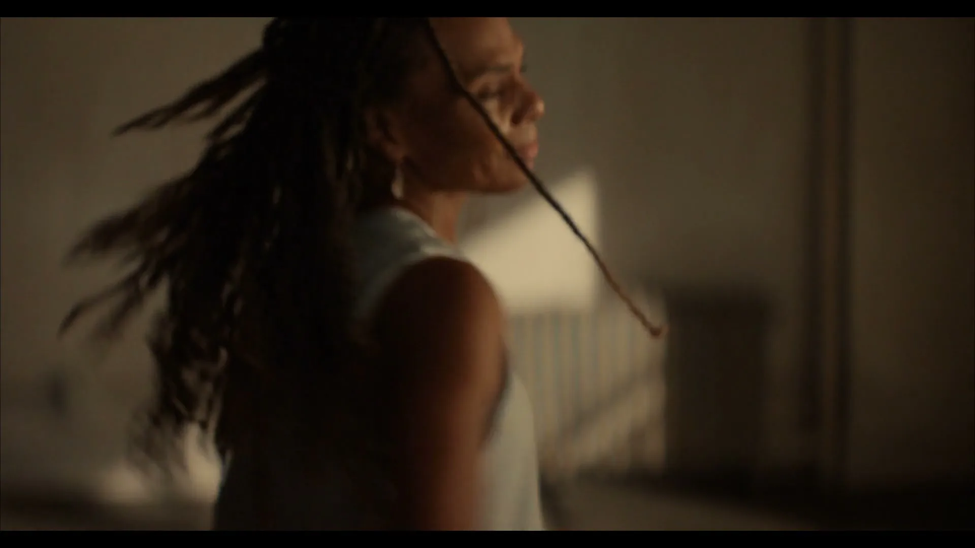A woman with dreadlocks in a light blue tank top dances, spinning around in a room with soft light and shadows.