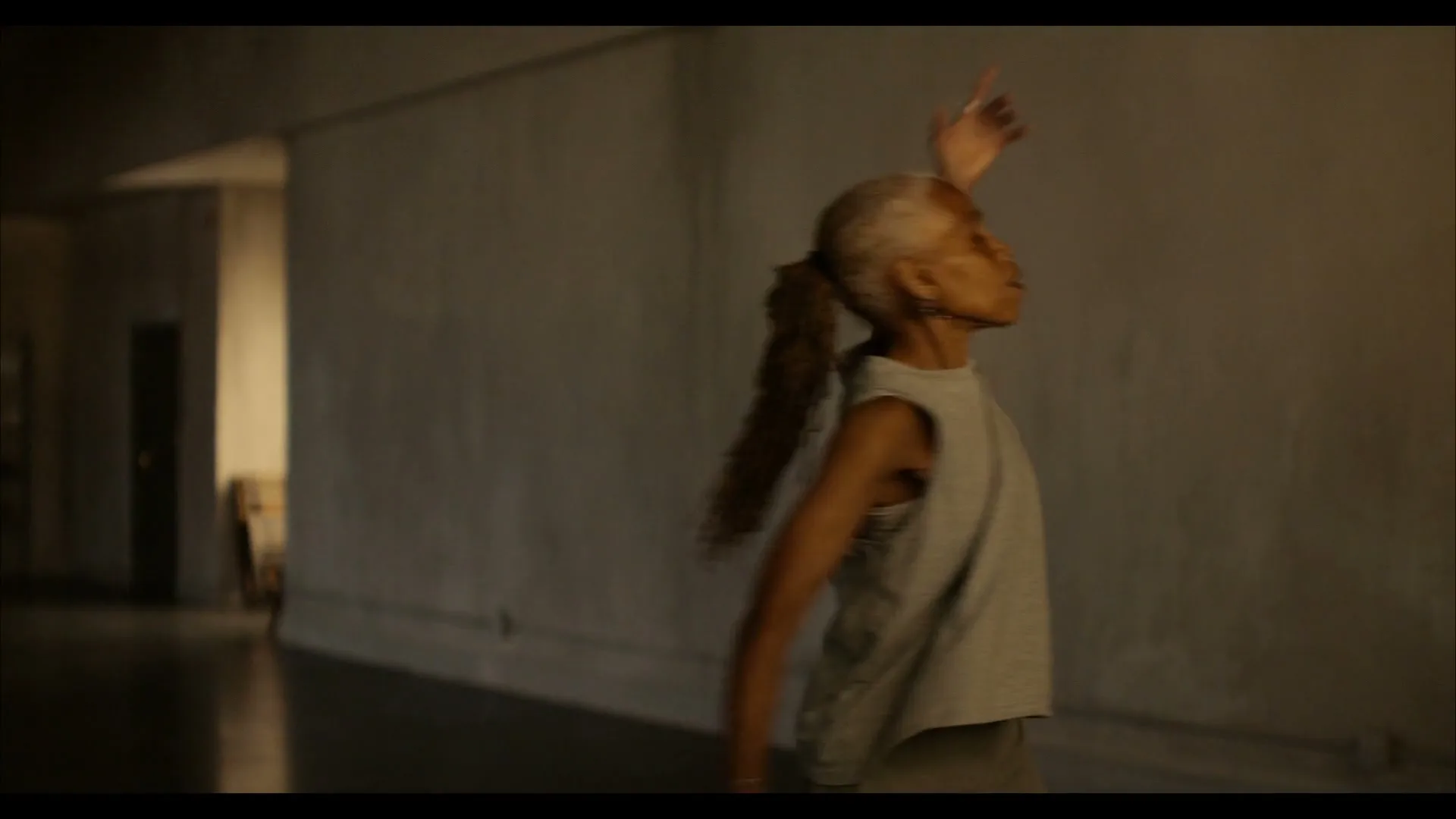 A woman with dreadlocks in a tank top and pants dances gracefully in a dimly lit, empty industrial space, arms outstretched.
