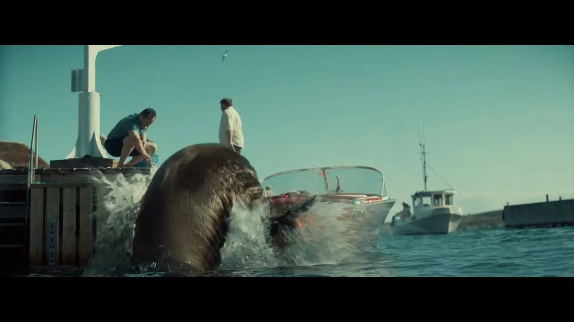 A walrus emerges from the water next to a wooden boat, surprising two men on a dock on a sunny day.