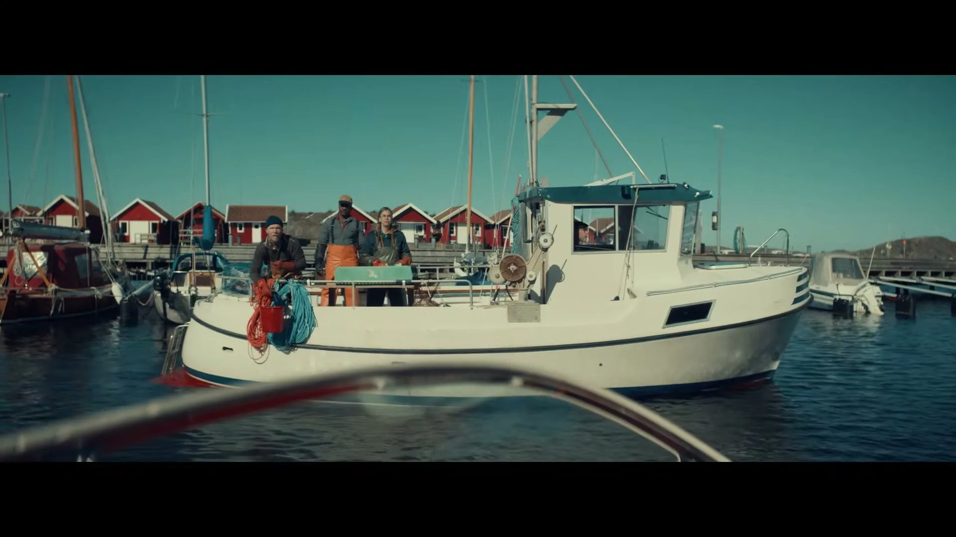Three fishermen stand on a white boat in a harbor with red boathouses in the background on a sunny day.