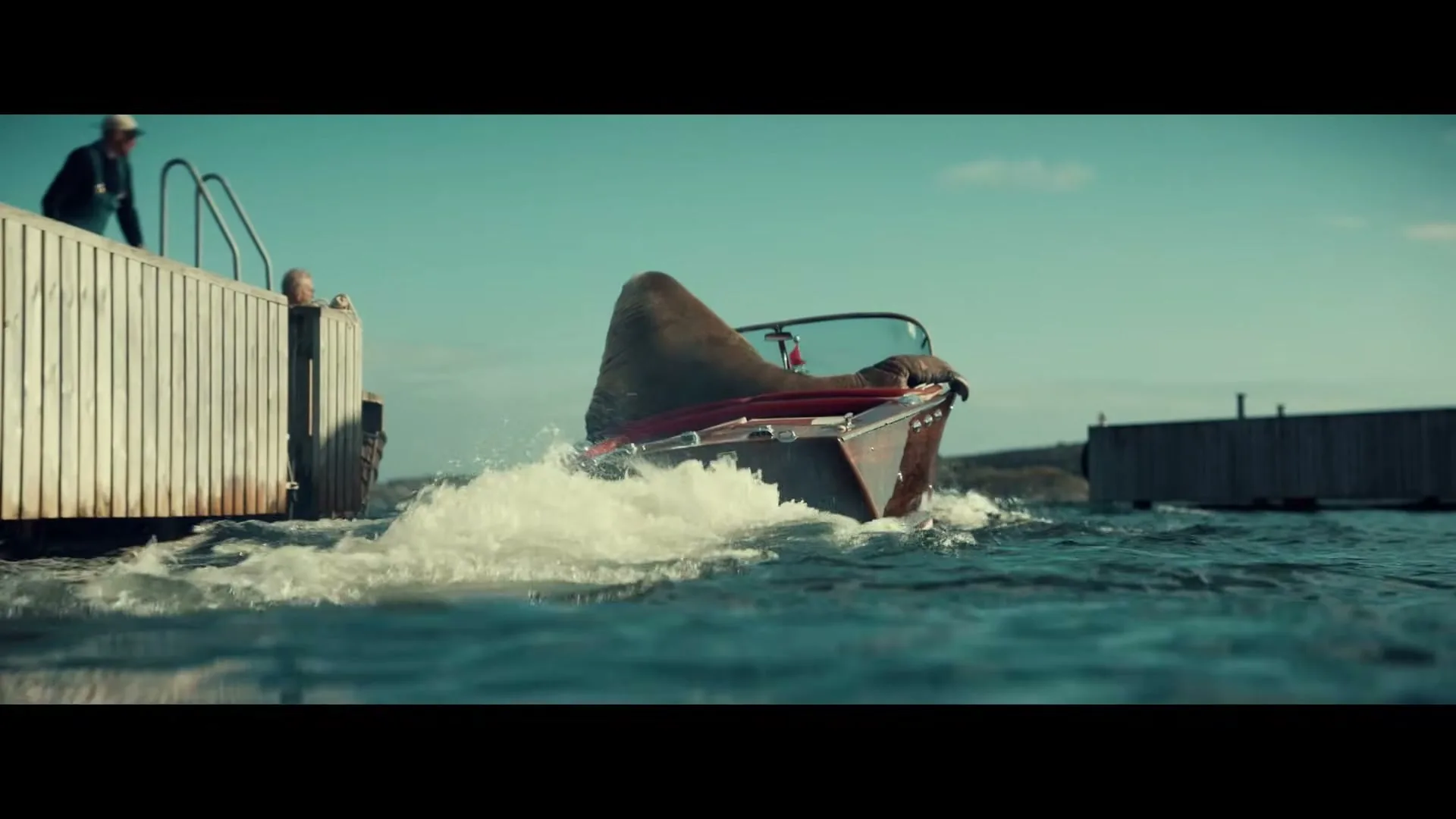 A walrus in a red speedboat speeds away from a wooden dock on a sunny day, creating a large wake in the blue water.