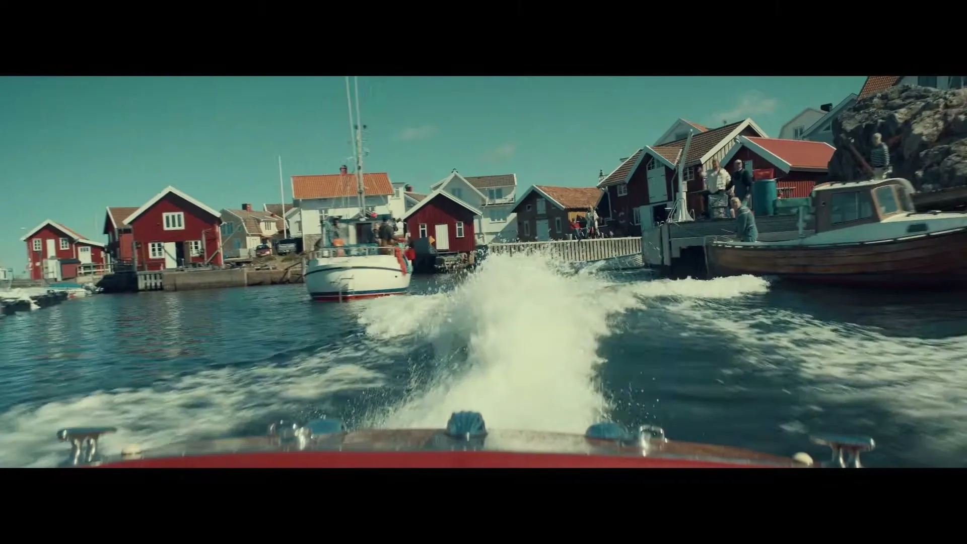 A red speedboat speeds away from a small harbor town with colorful houses and boats, leaving a white wake behind it on a sunny day.