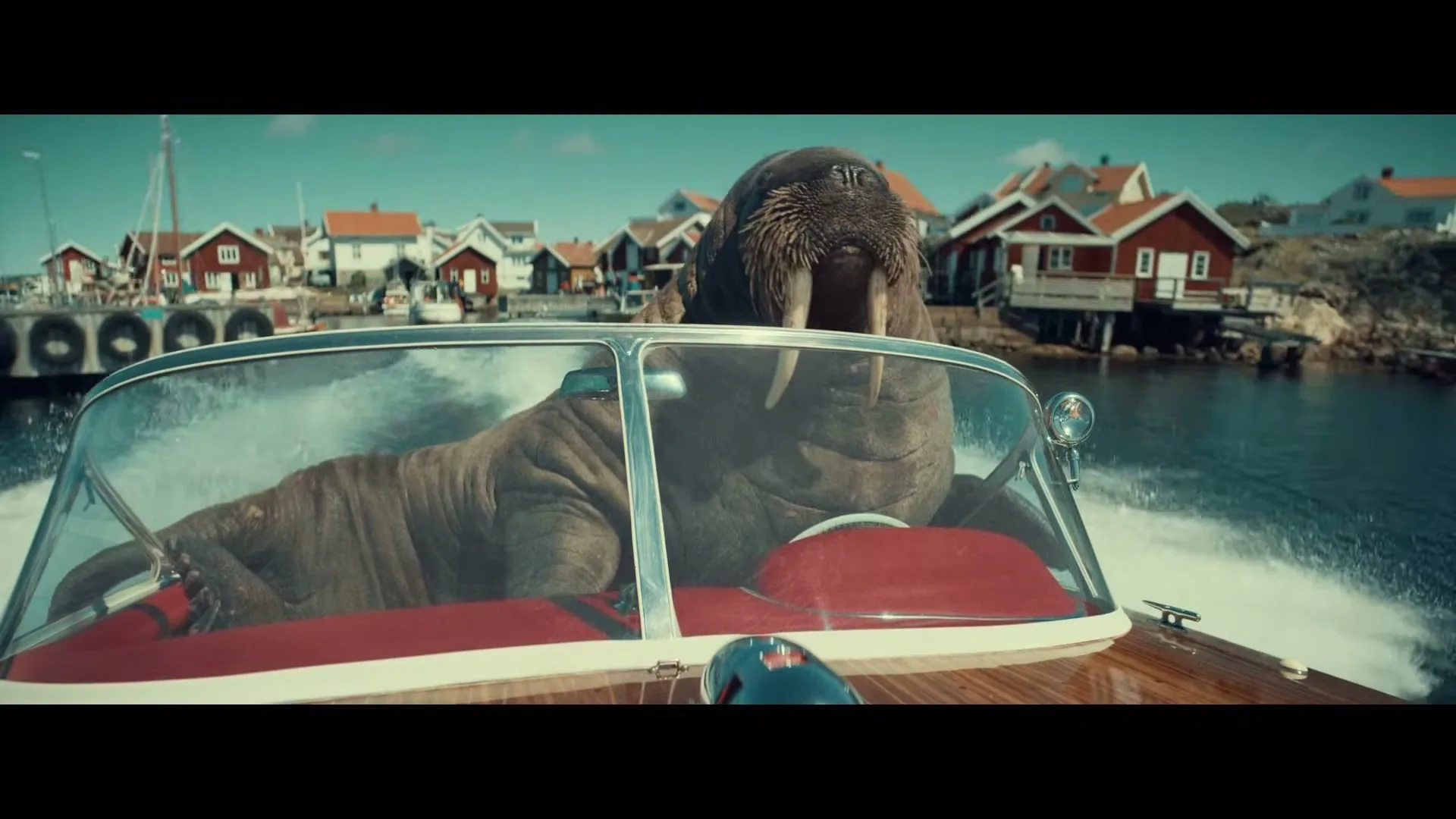 A walrus in a speedboat drives past a small coastal village with red houses under a clear blue sky.