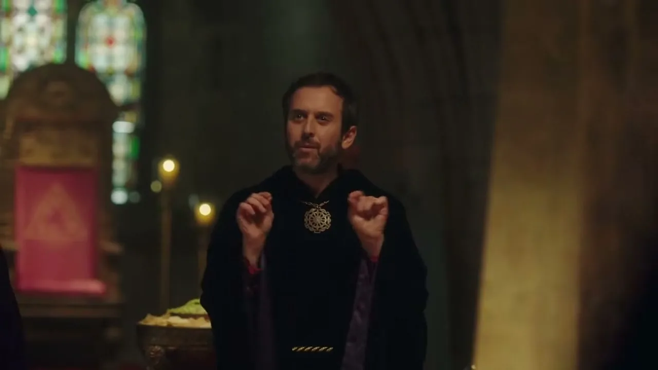 A man with a beard, wearing a black robe and a medallion, gestures with his hands while speaking in a dimly lit, ornate room with stained glass windows.