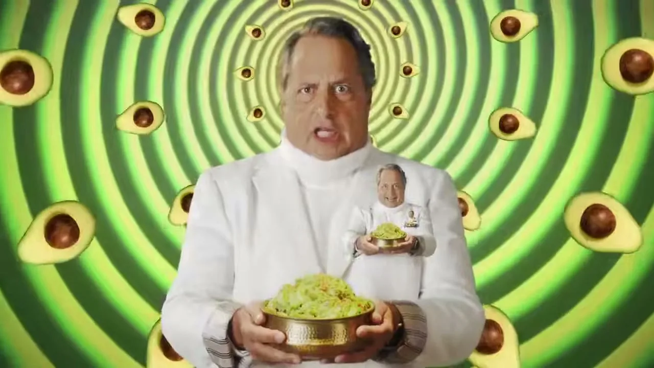A man in a white suit holds a bowl of guacamole, with a smaller version of himself on his chest, against a green swirling background with avocados.