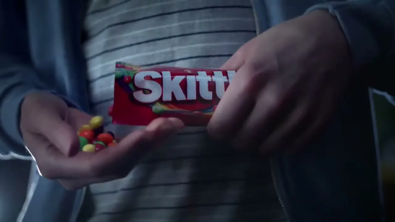 A person in a striped shirt and blue jacket pours Skittles from a red bag into their hand, in an interior setting.