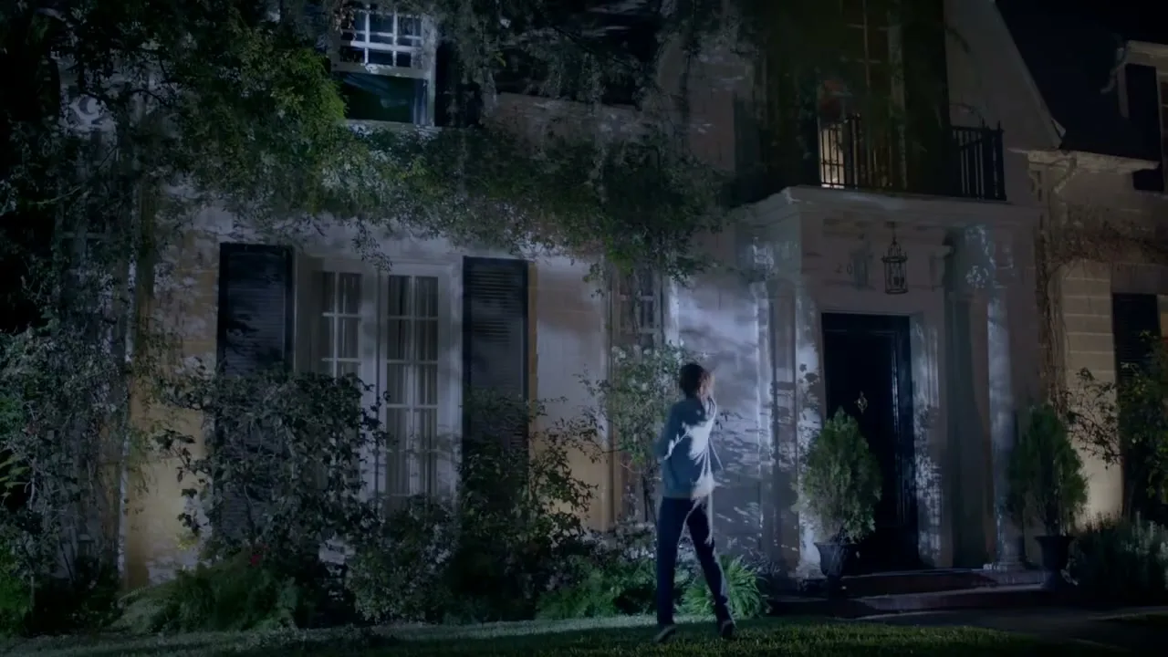 A young man in a hoodie stands on a lawn at night, looking up at a large, ivy-covered house with illuminated windows, calling out a name.