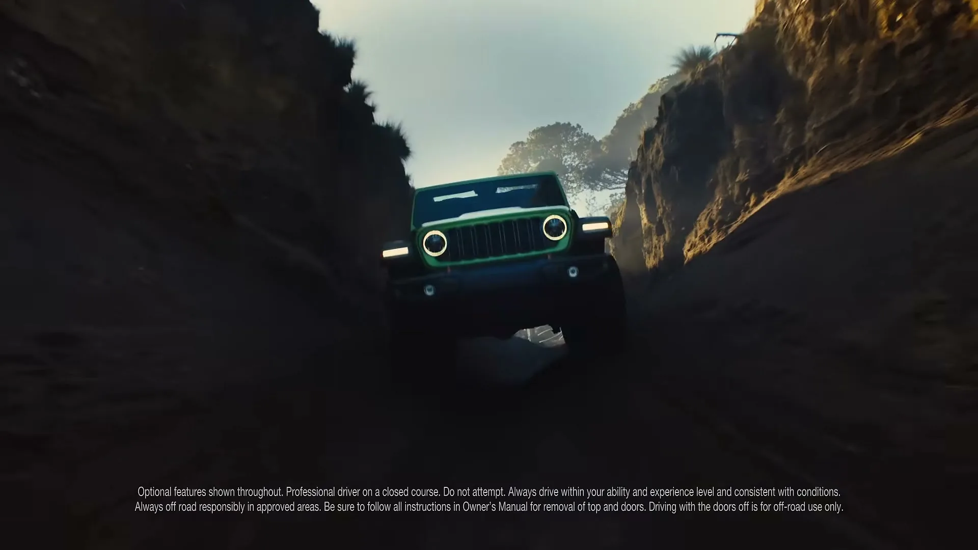A green Jeep Wrangler drives towards the camera on a dirt path between two rocky cliffs under a bright sky.