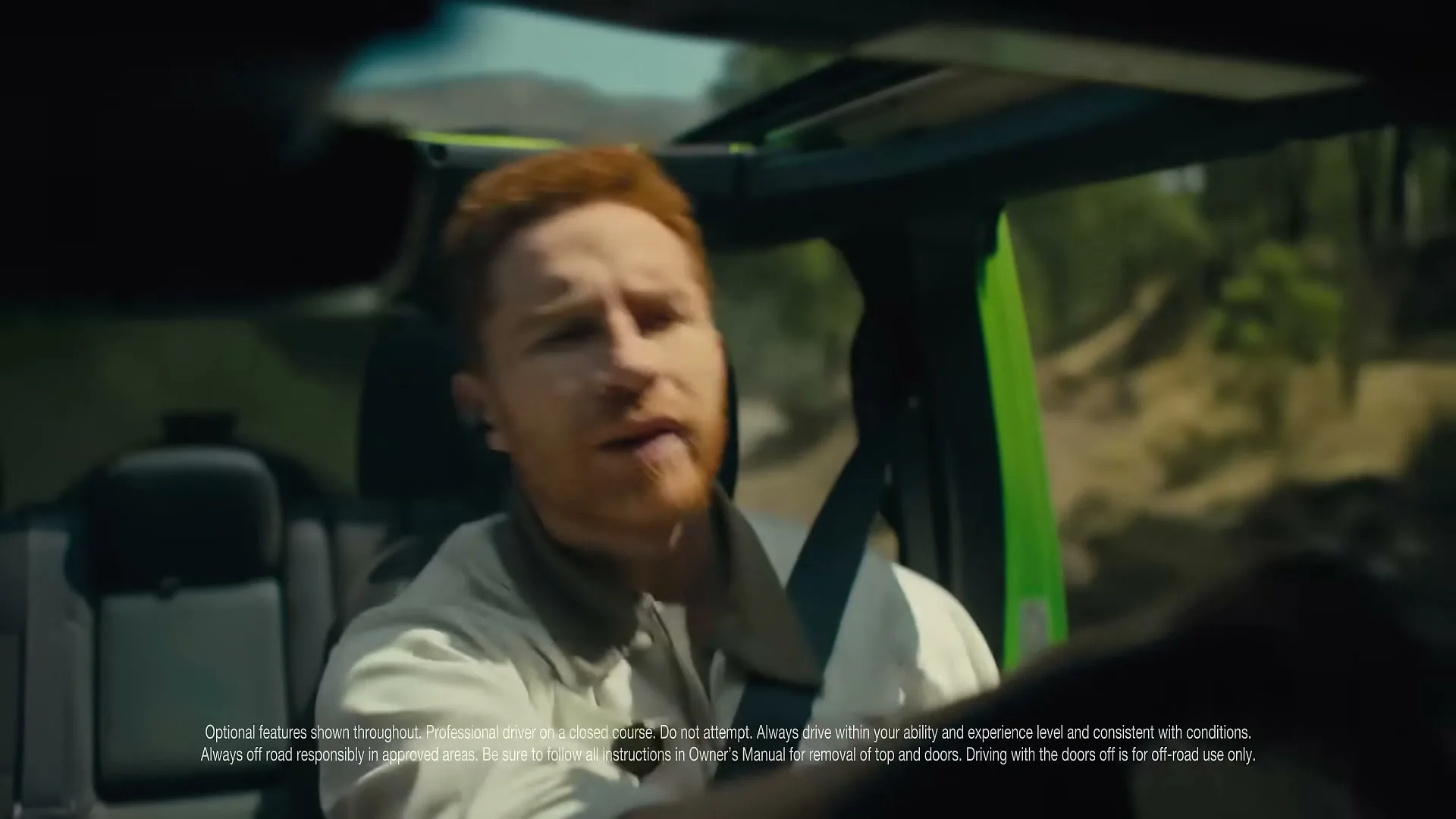 A red-haired man in a light jacket drives a green Jeep on a dirt road, looking surprised and talking.