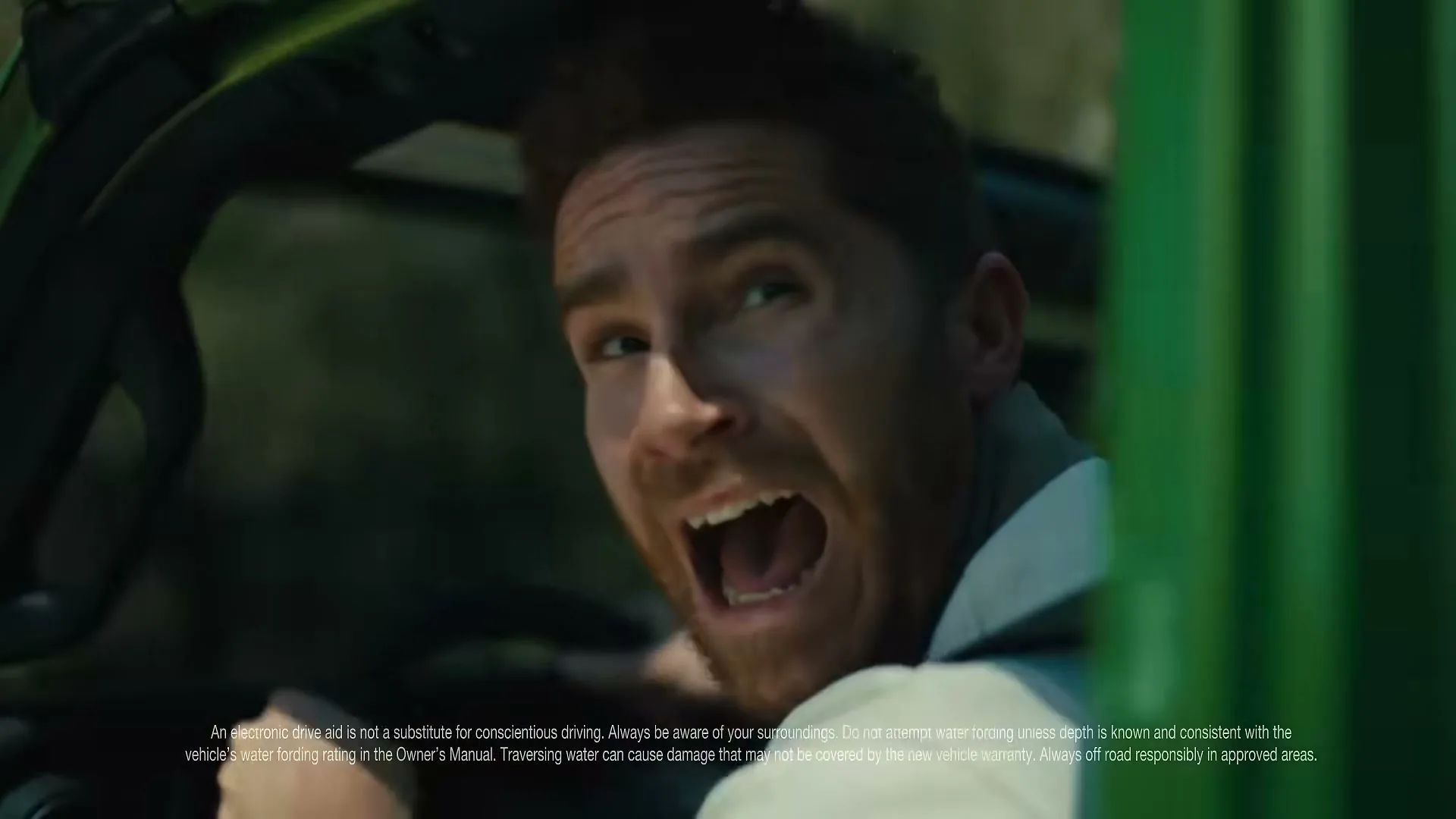 A man with a beard and white shirt looks back in fear from inside a green vehicle.