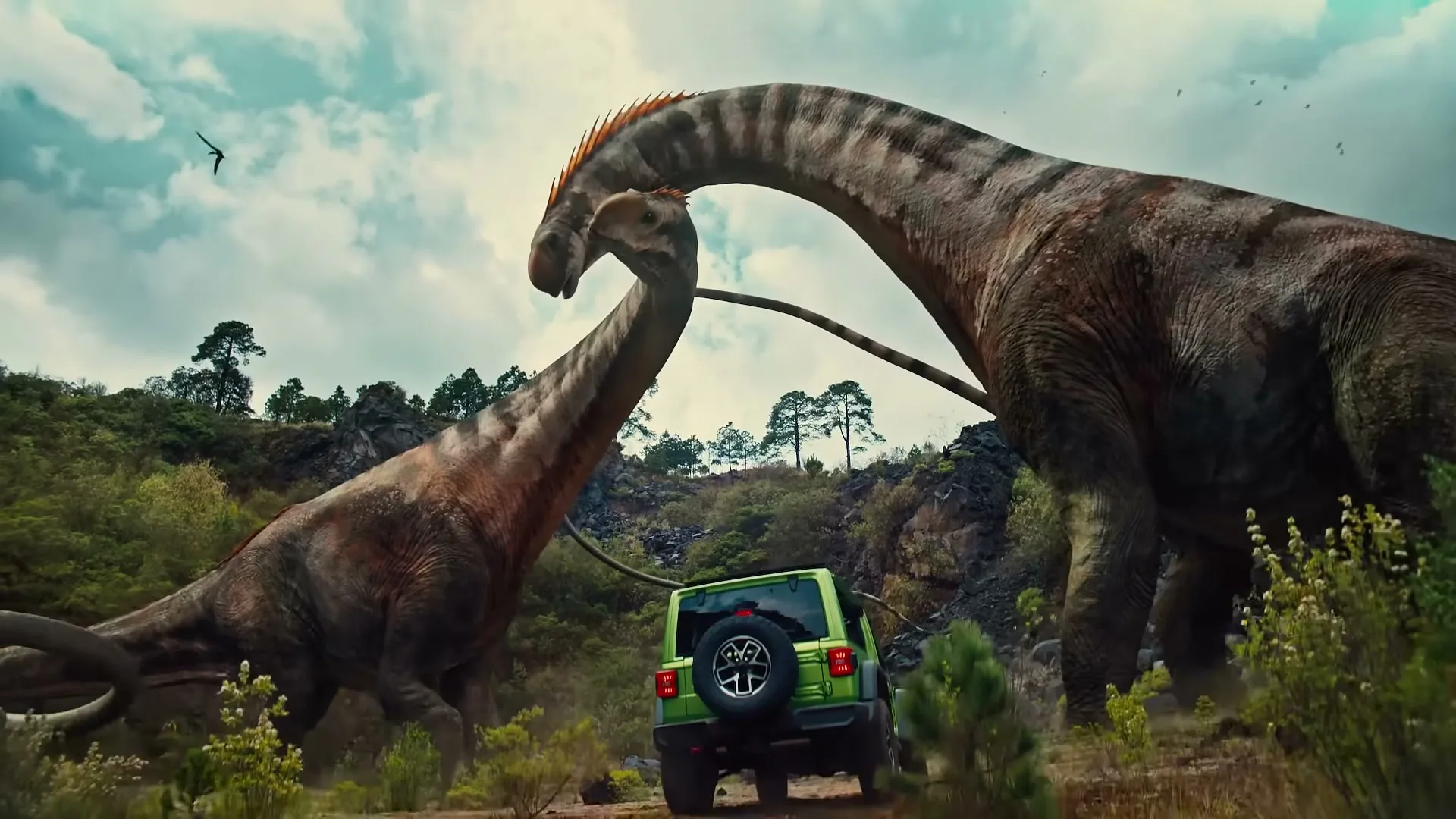A green Jeep Wrangler is dwarfed by two massive long-necked dinosaurs on a dirt road in a prehistoric landscape under a cloudy sky.