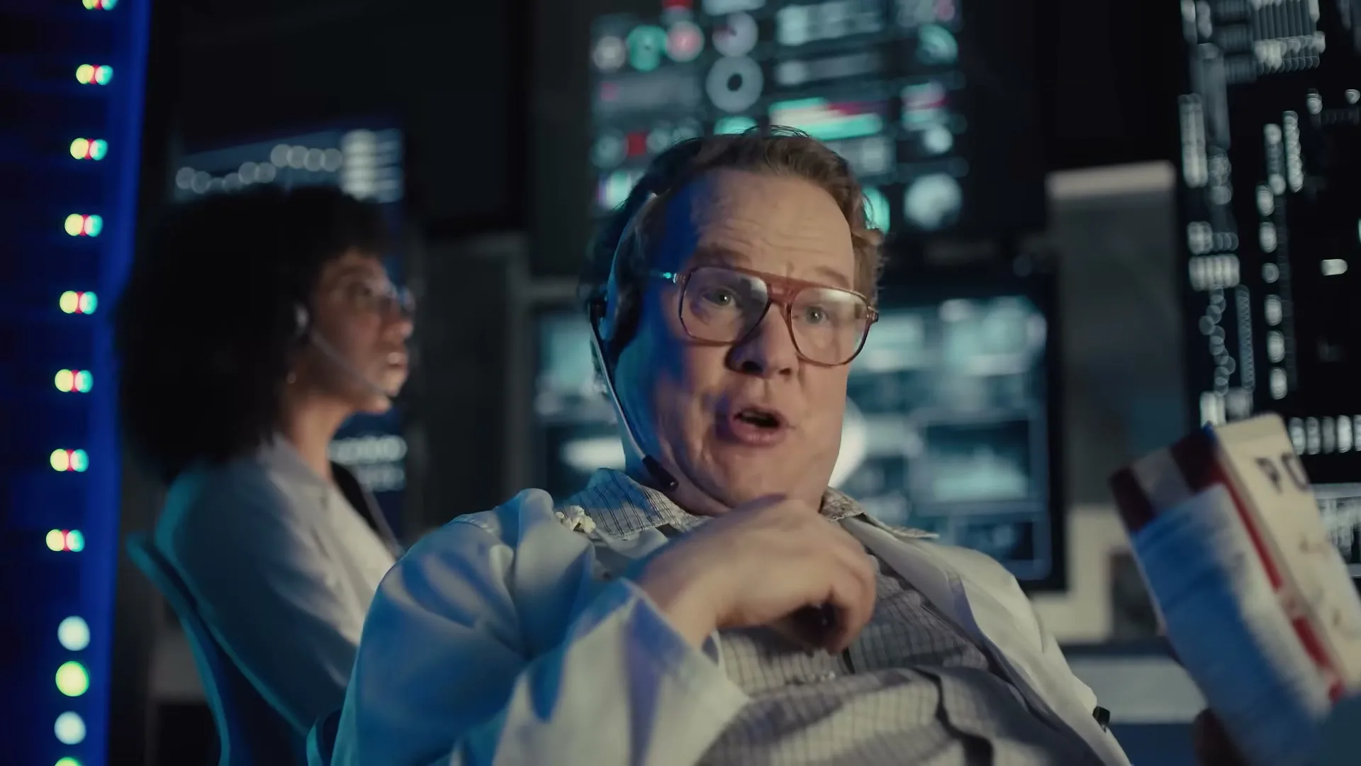 A male scientist in a lab coat and glasses looks surprised while a female scientist works in the background in a control room.