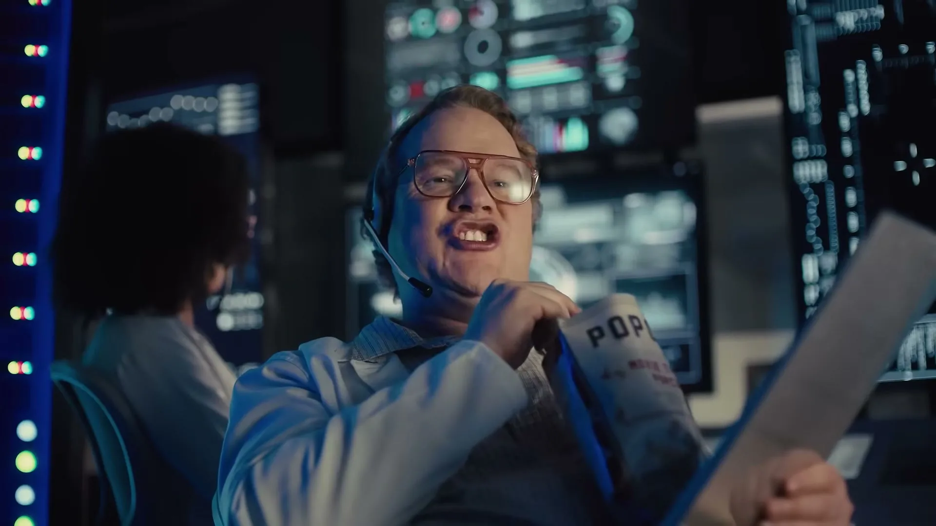 A male scientist in a lab coat and headset smiles while eating popcorn and holding papers in a dark control room with multiple screens.