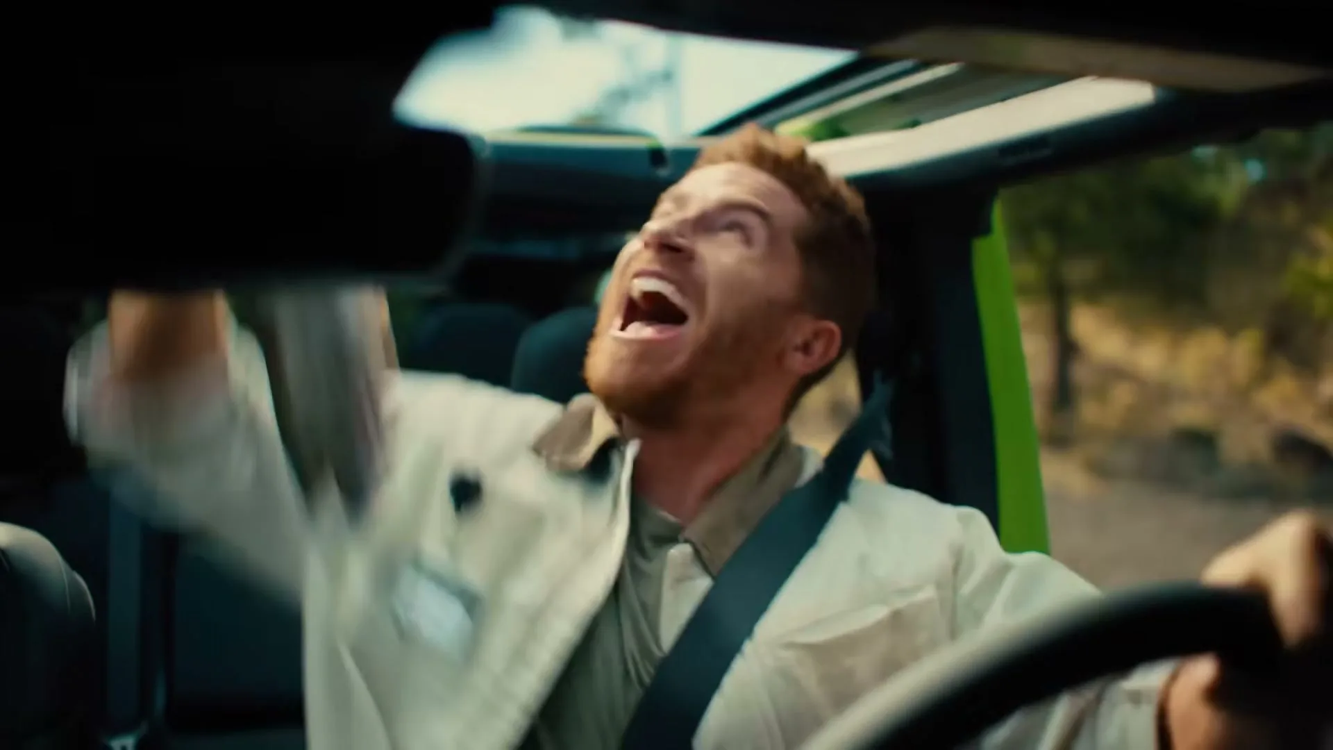 A man with red hair and a beard, wearing a light jacket, screams in terror while driving a green Jeep with the top down through a forest.