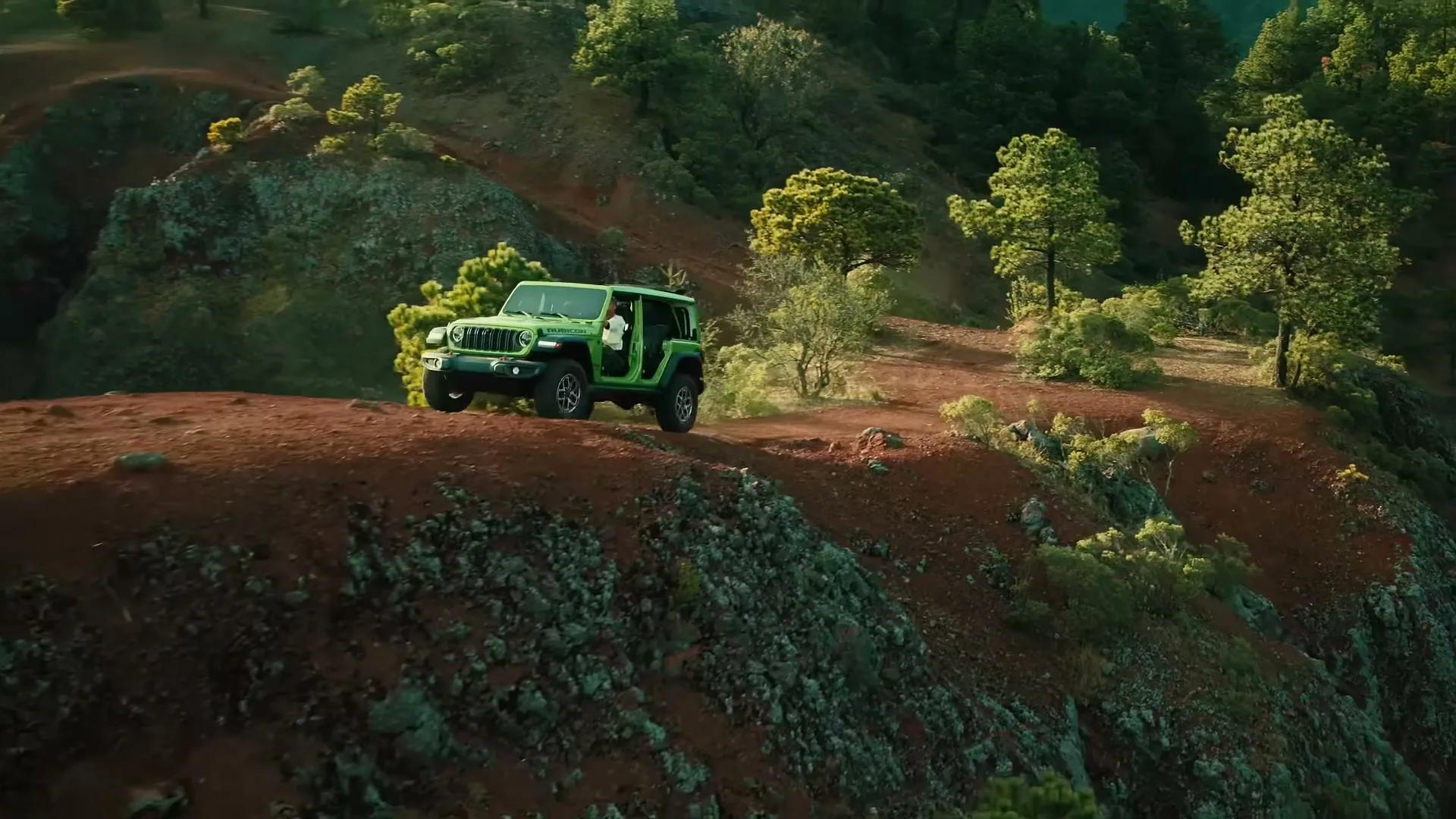 A green Jeep Wrangler drives along a narrow, winding dirt road on a steep, forested mountainside during the day.
