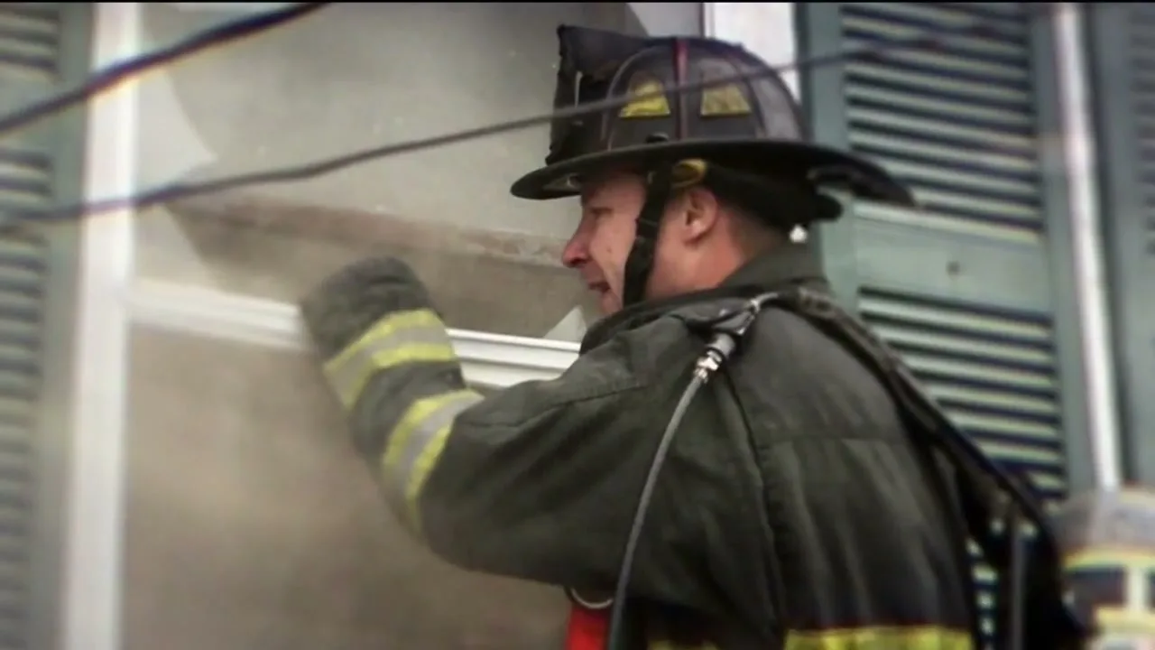 A male firefighter in full gear sprays water into a smoky window of a building, with a focused and determined expression.