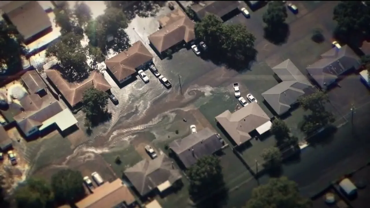 An aerial view shows a suburban neighborhood with houses and cars partially submerged in floodwaters, with swirling patterns visible in the murky water.
