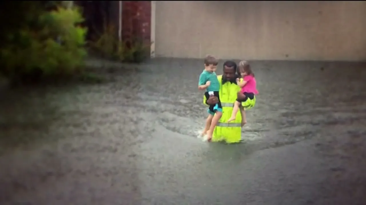 A man in a bright yellow vest carries two young children through a flooded street, with buildings and trees in the background.