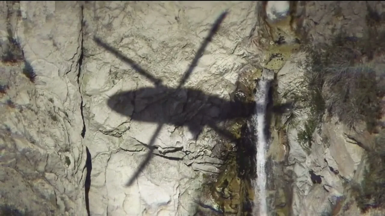 A helicopter shadow is cast on a rocky cliff face with a small waterfall, as the camera looks down from above.