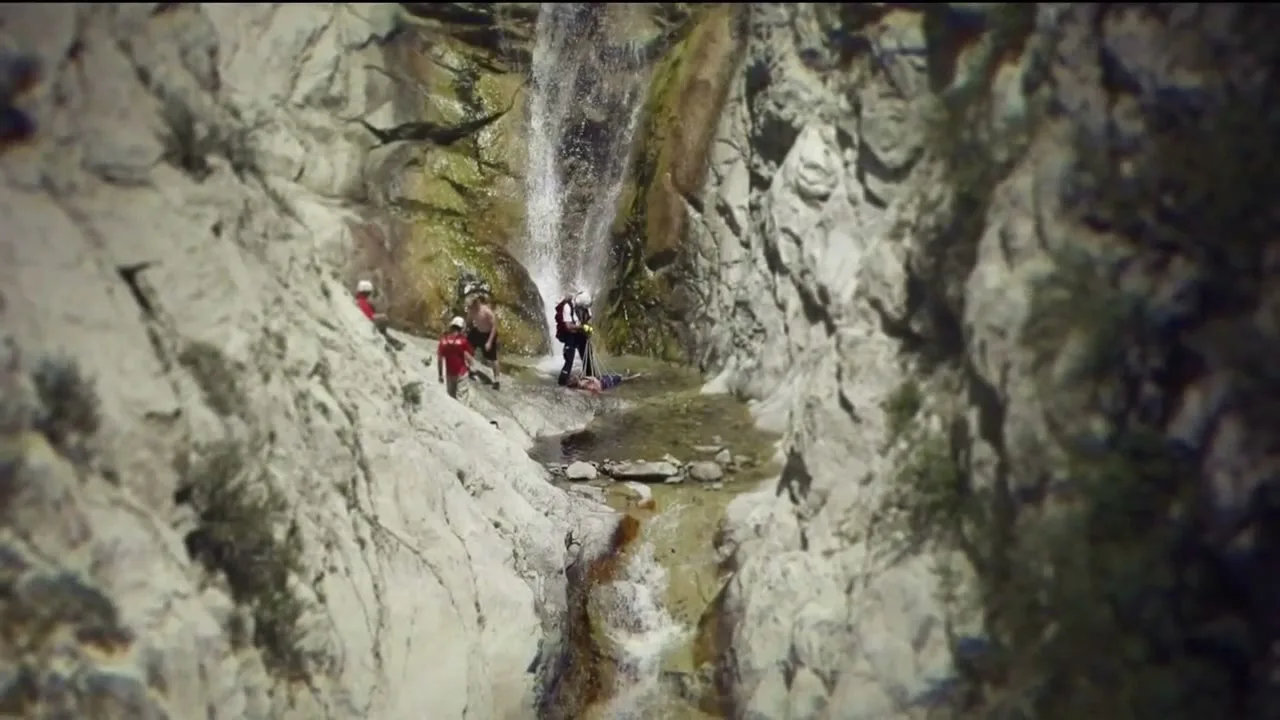 A group of rescue workers in helmets and safety gear attend to an injured person lying on the ground near a waterfall in a rocky canyon.