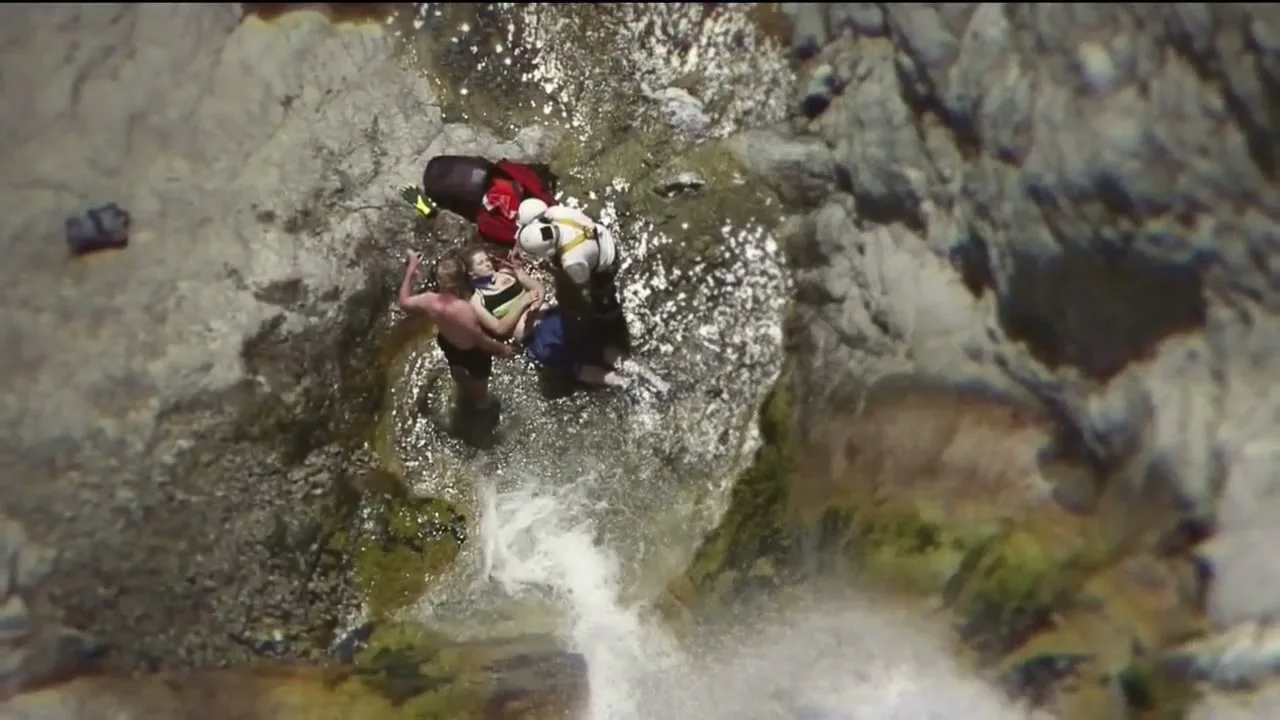 An aerial view shows a rescue worker in a white helmet and red backpack helping a person in a life vest in a rocky river with a waterfall.