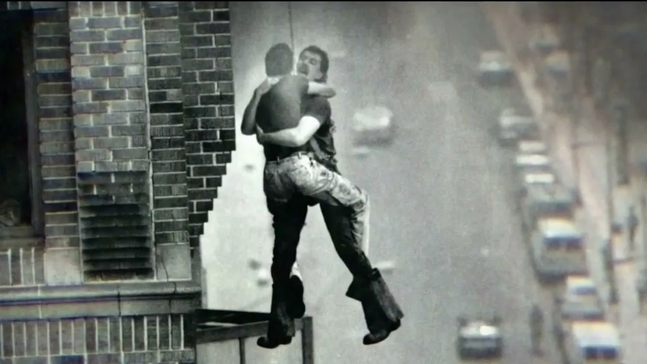 Two men embrace while one hangs from a rope, with a city street and buildings in the background, in a black and white archival photo.