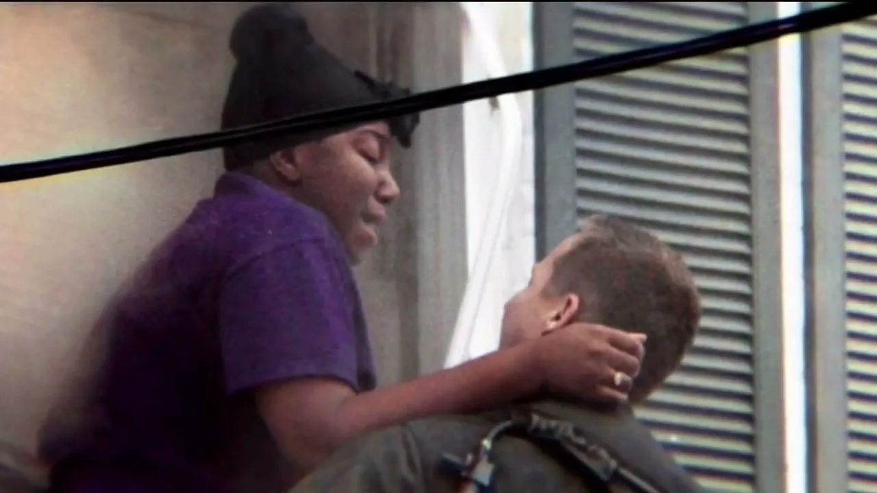A woman in a purple shirt and black beanie embraces a man in a dark uniform, looking at him with a pained expression in a building opening.