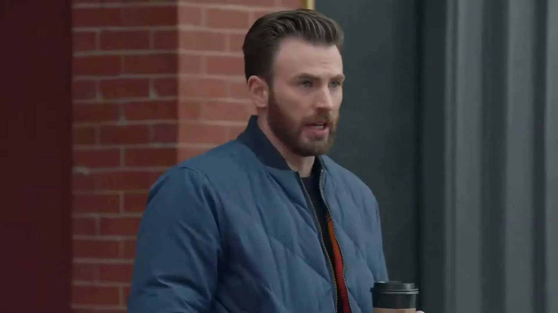 A man with a beard and blue jacket holds a coffee cup and looks to the side, standing outside a brick building.