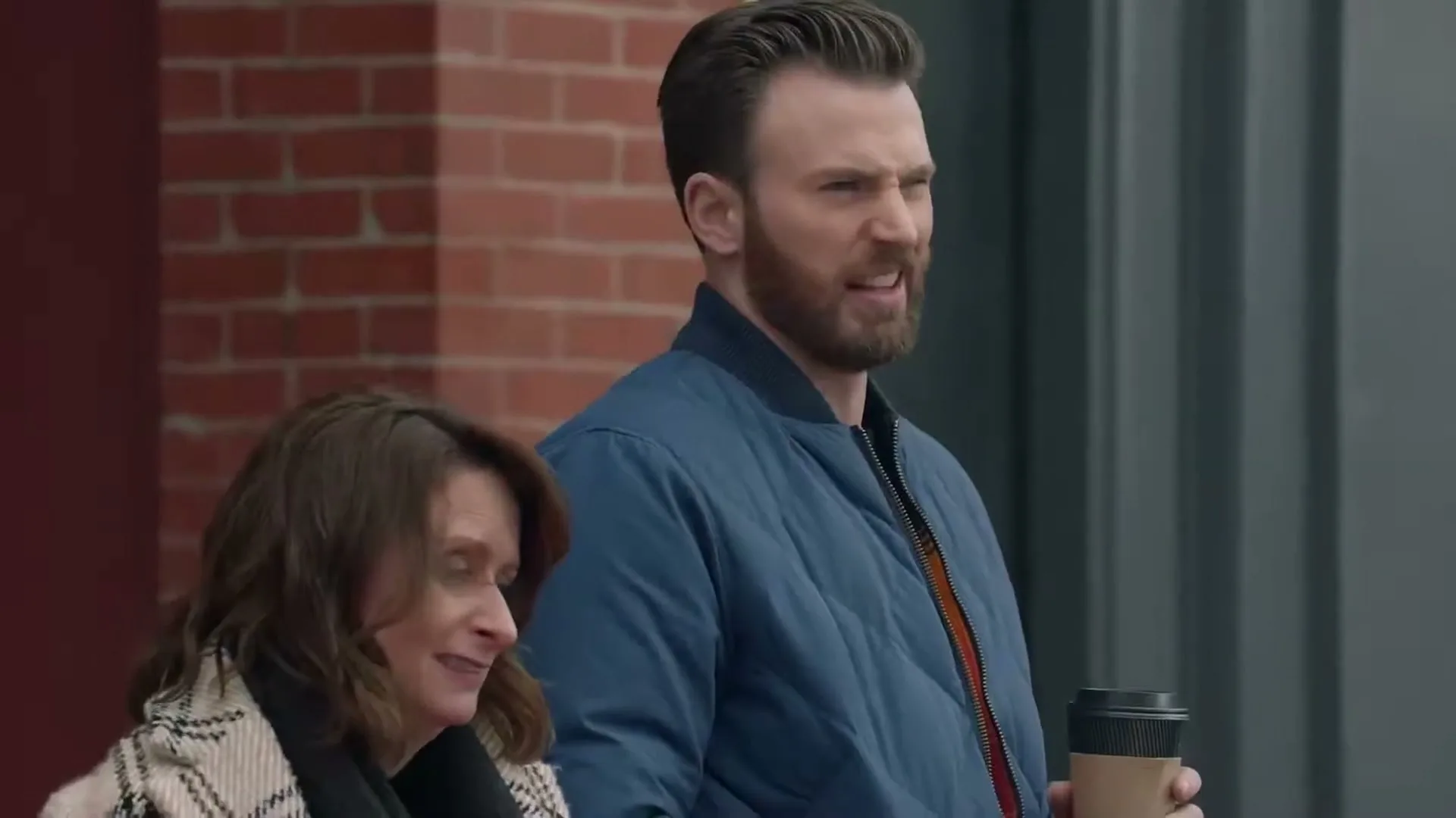 A man with a beard and a woman with brown hair stand outside a brick building, looking off-camera with expressions of skepticism and amusement.