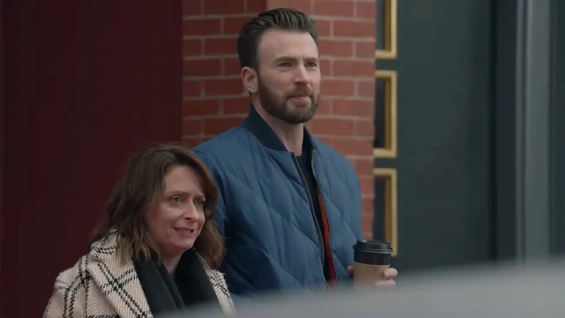 A man and a woman stand on a sidewalk, looking off-screen with curious expressions, as the woman speaks in a Boston accent.