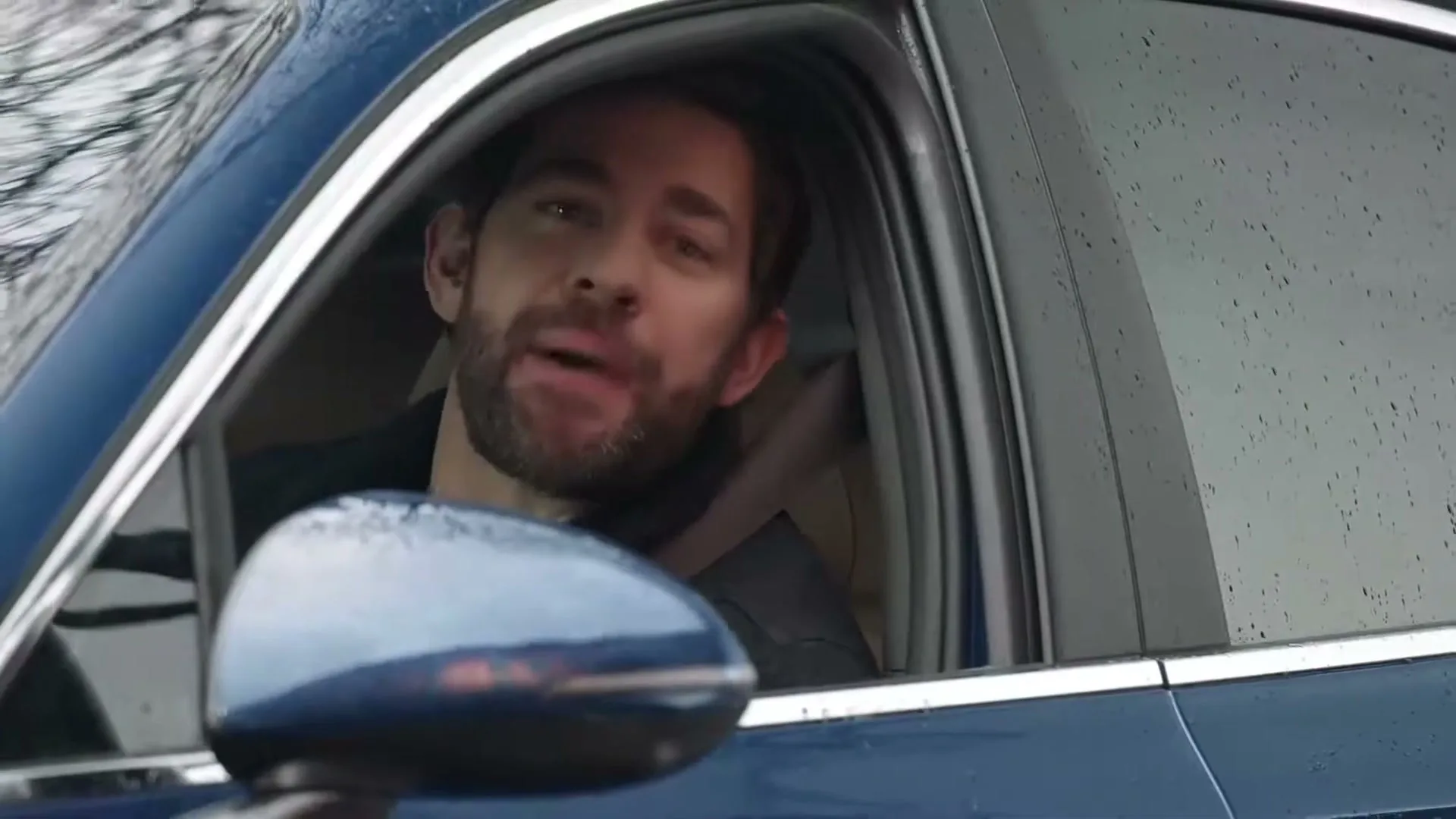 A man with a beard, wearing a dark jacket, winks and smiles from the driver's seat of a blue car on a rainy day.