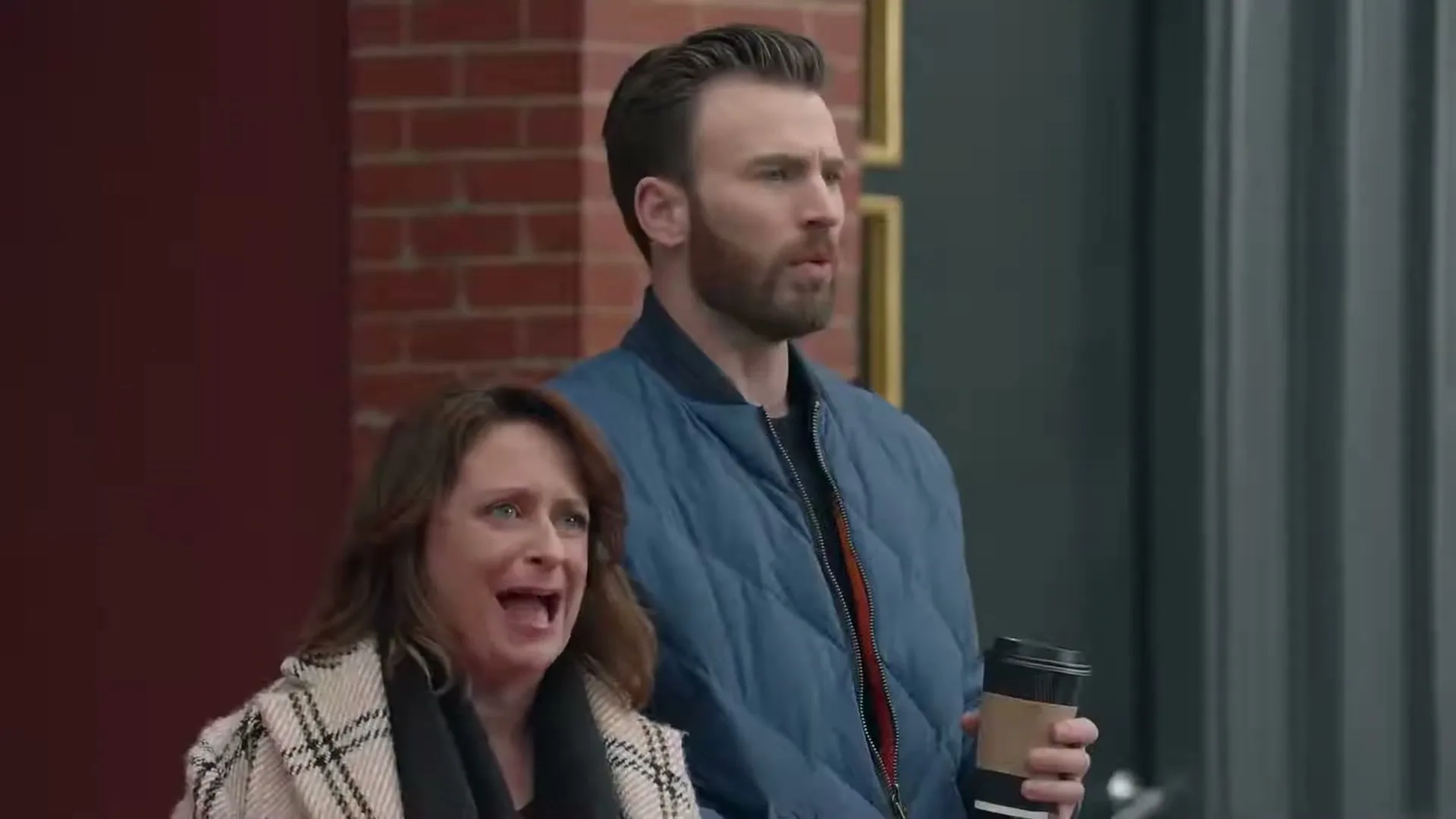 A woman in a plaid coat and a man in a blue jacket stand outside a brick building, looking surprised and talking.
