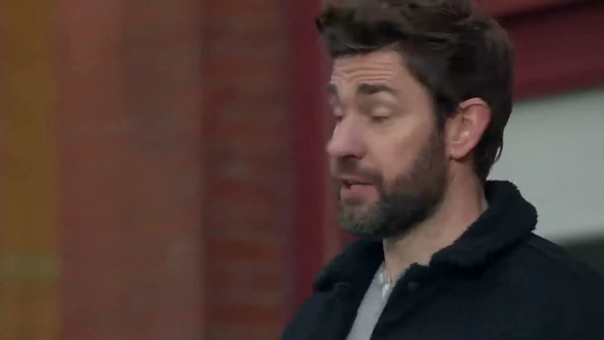 A man with a beard and dark jacket looks off-camera, speaking in a suburban setting during the day.
