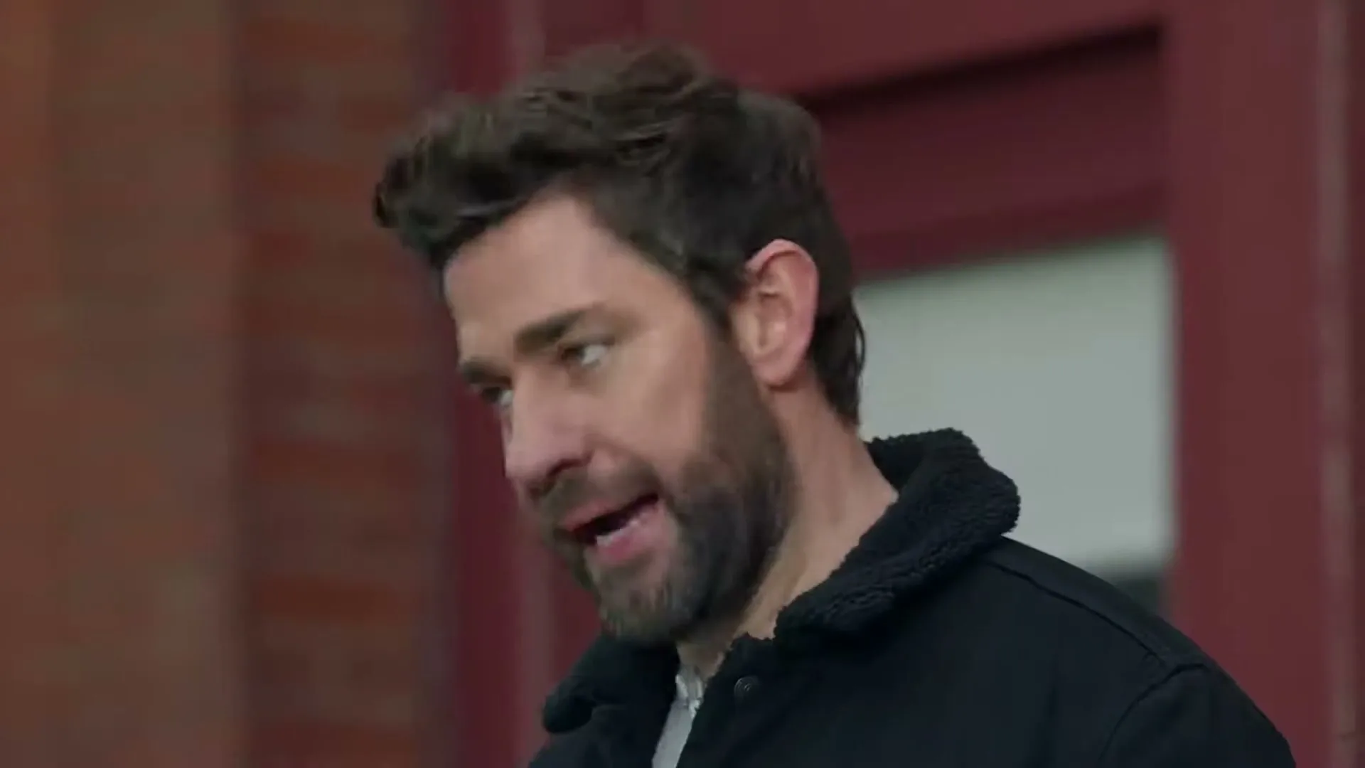 A man with a beard and dark jacket looks down, then up and to the side, speaking in an urban exterior.