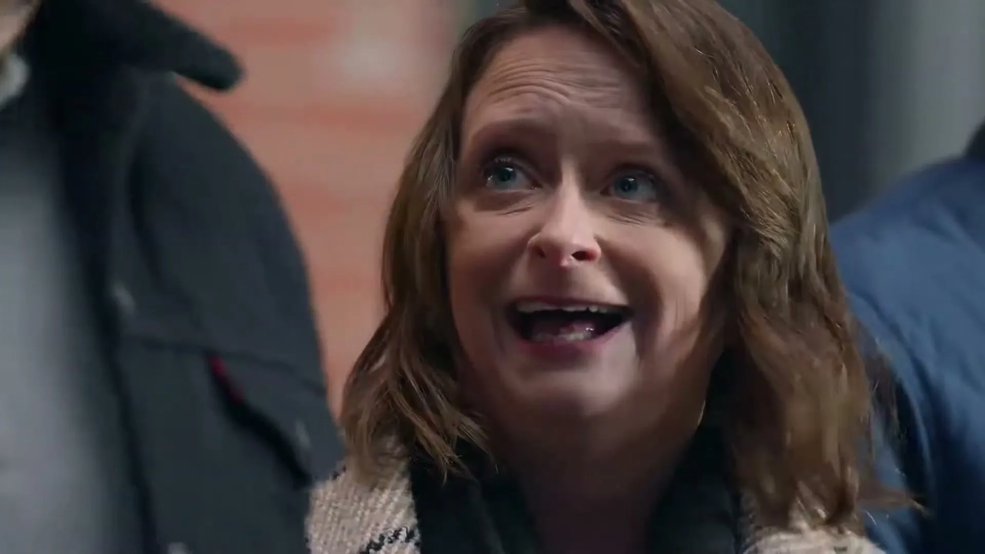 A woman with brown hair and a patterned coat looks surprised and speaks to someone off-screen in an urban setting.