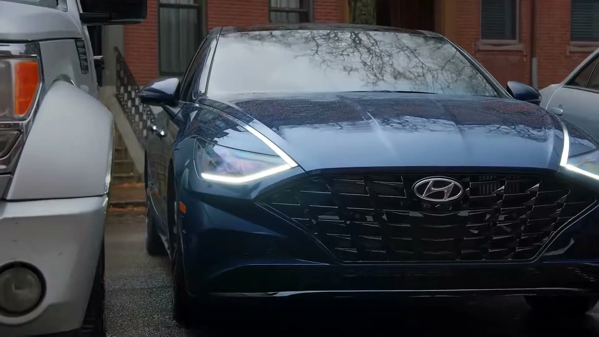 A blue Hyundai Sonata with its headlights on is parked between two other cars on a wet street in front of a brick building.