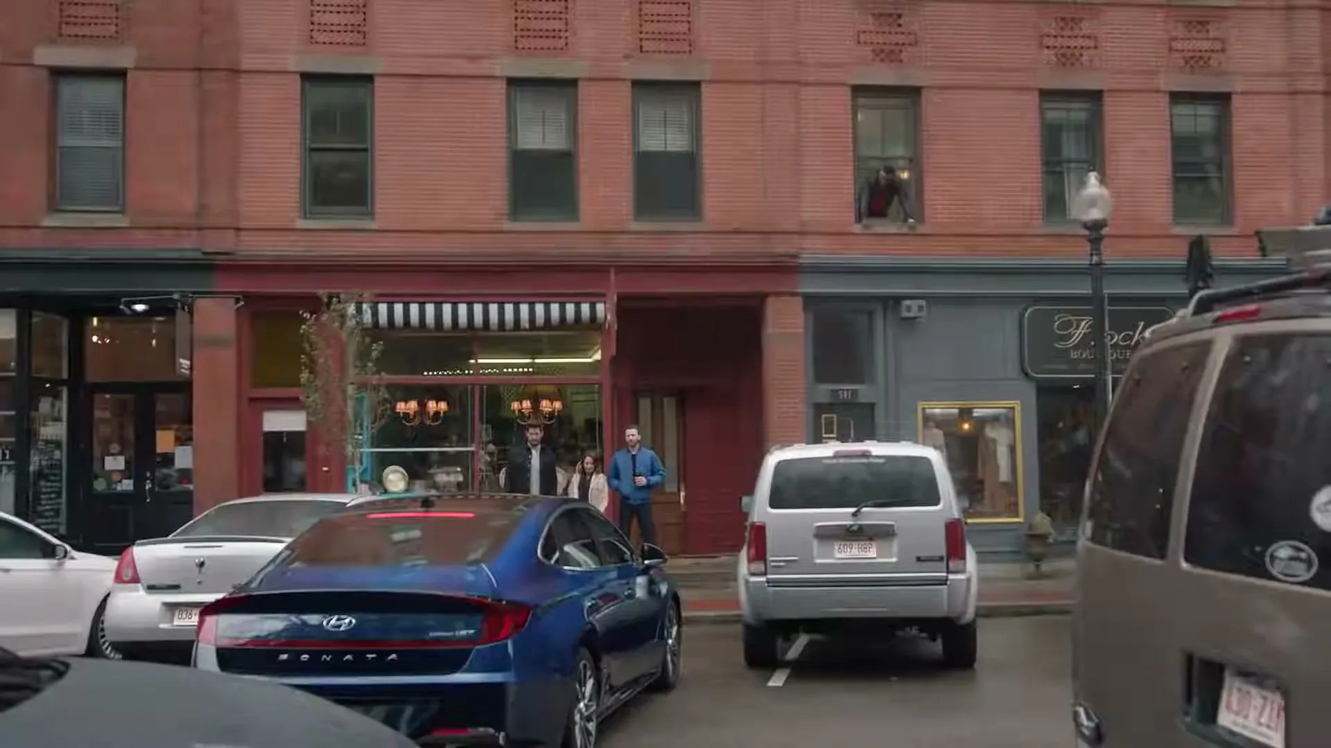 A blue Hyundai Sonata is parked on a city street, with people standing outside a storefront and a man looking out a window above.