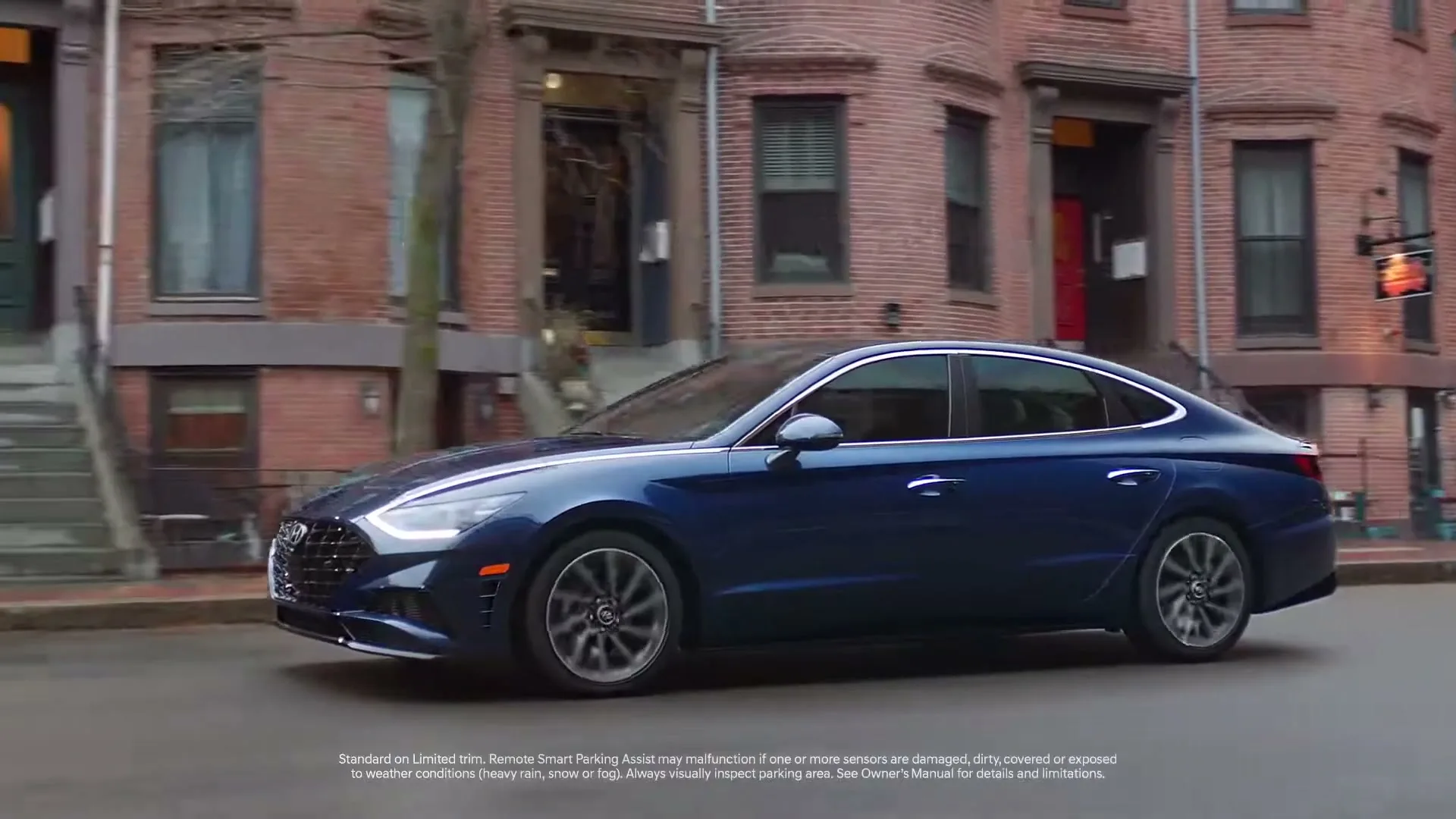 A dark blue Hyundai Sonata drives slowly on a city street, with brick buildings and bare trees in the background, as text appears on screen.